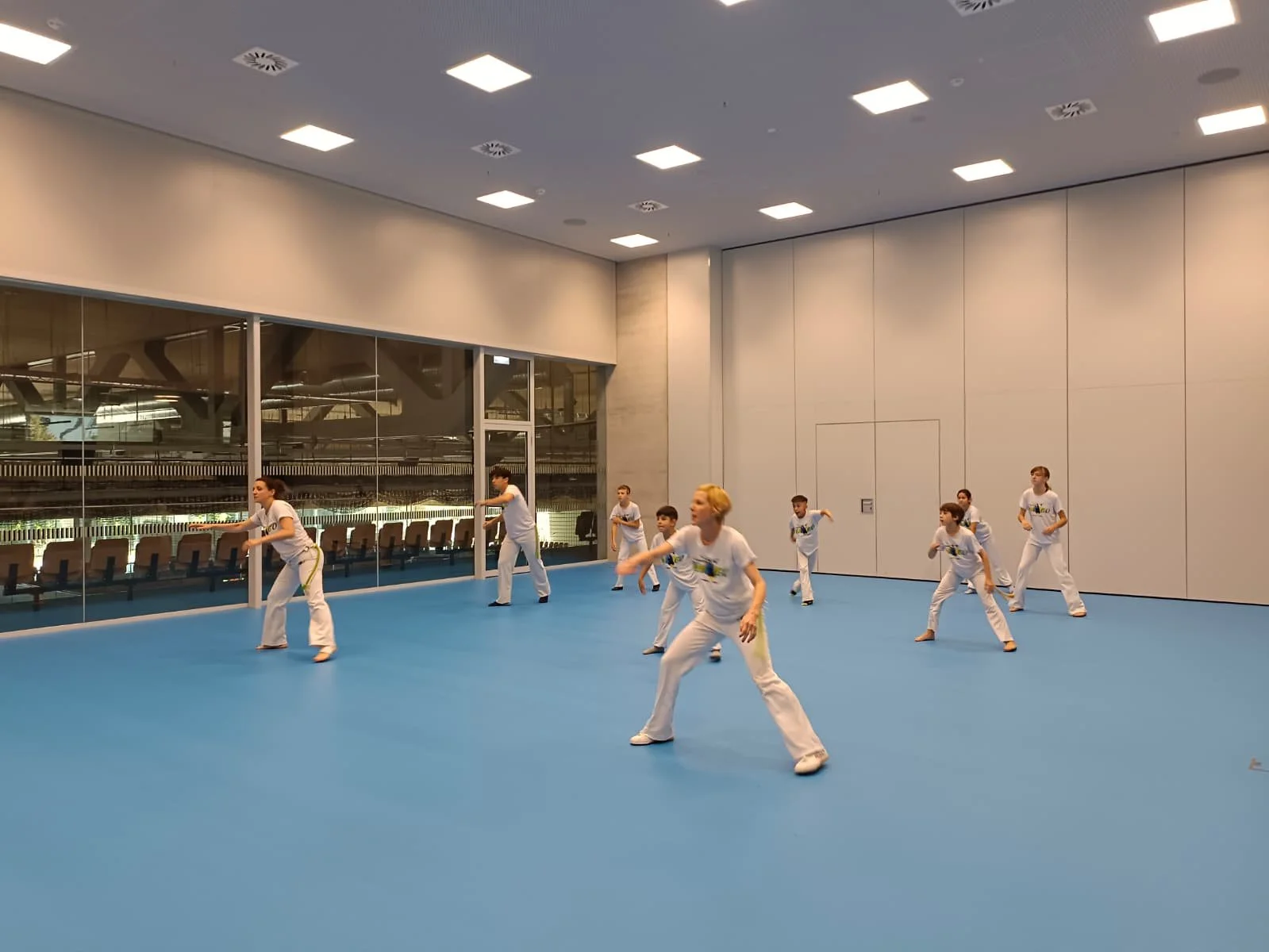 Kinder trainieren Capoeira in einem modernen Turnraum mit blauer Matte und großen Fenstern.