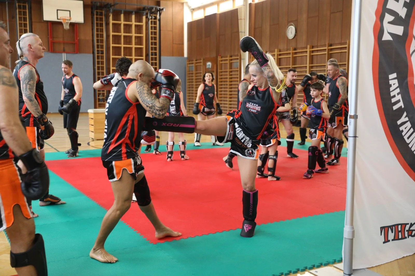 Muay Thai Kampftraining in einer Sporthalle mit mehreren Kämpfern in Kampfanzügen, die in einer Trainingssession üben, inklusive Schlägen und Tritten, mit einem Trainer im Hintergrund.