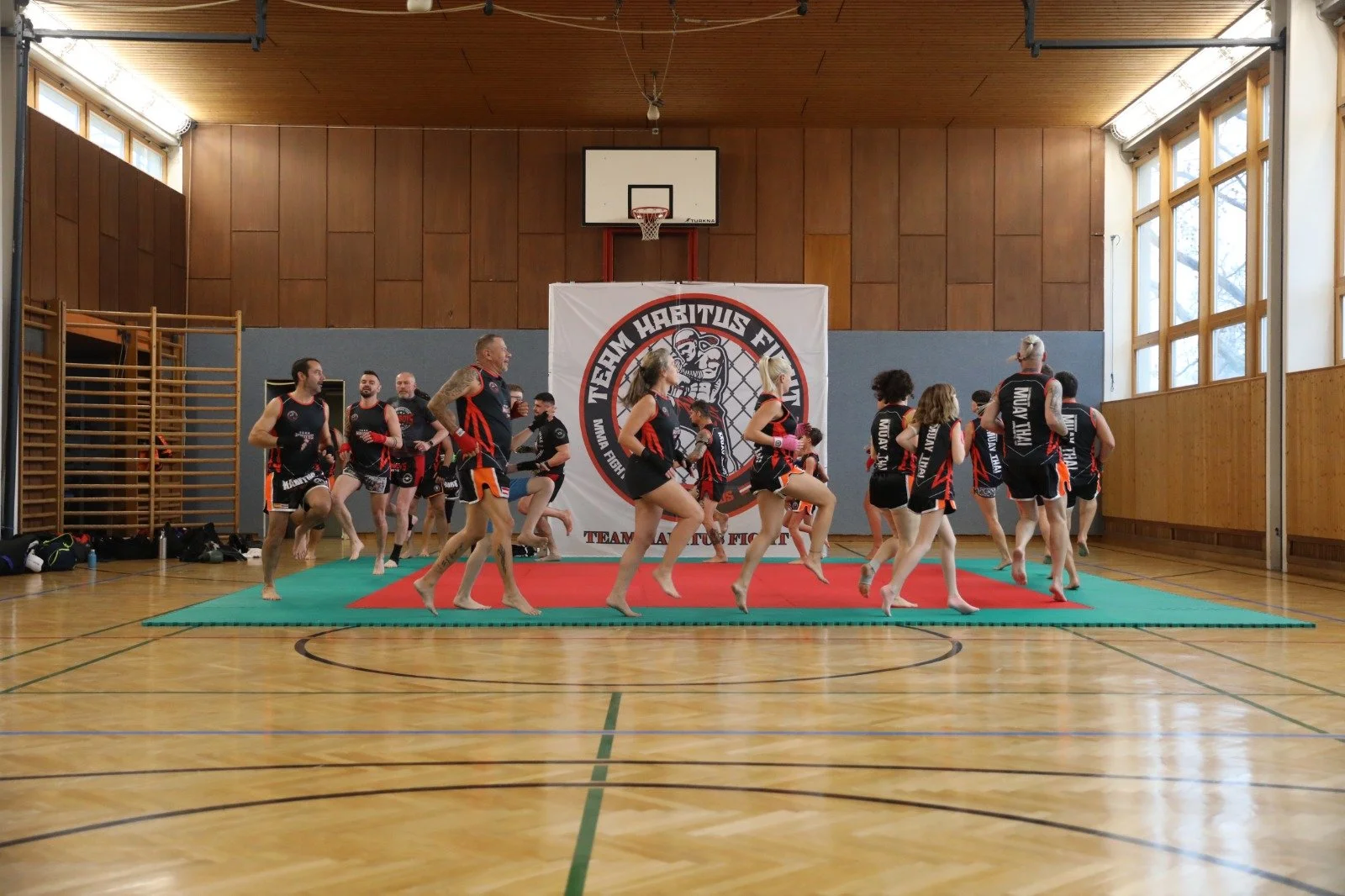 Gruppentraining im Fitnessstudio mit Frauen und Männern in schwarzen und roten Sportkleidern, die auf einer Matte im Turnraum üben, mit einem Banner im Hintergrund, das den Namen eines Kampfsportteams zeigt.
