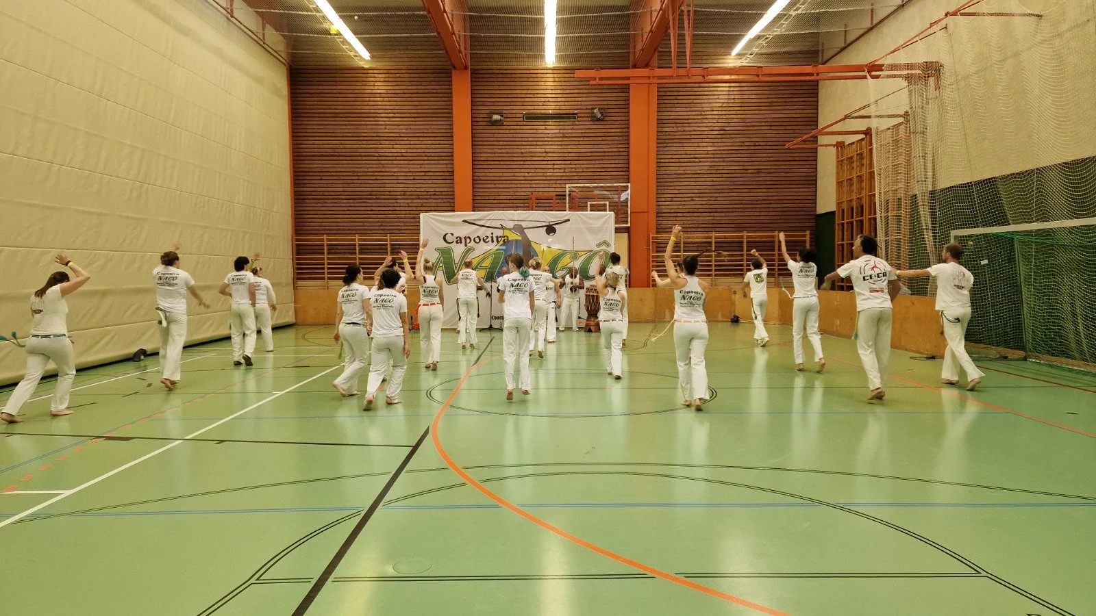 Gruppe von Menschen, die Capoeira im Fitnessstudio üben, trägt weiße Kleidung mit Capoeira-Prints, auf einer grünen Sporthalle mit Ballett- und Turngeräten.
