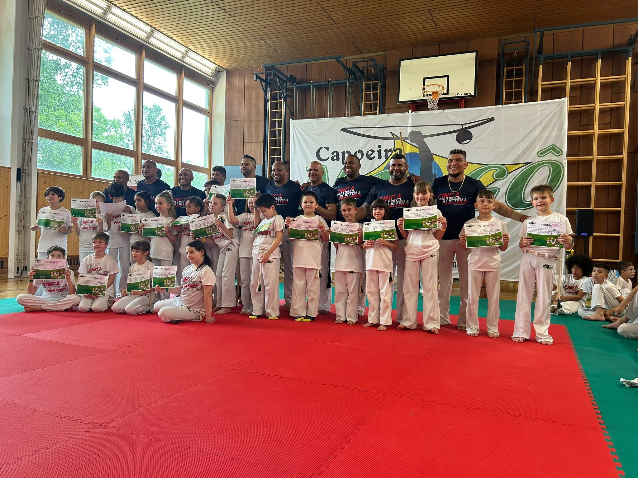 Gruppenfotografie bei einer Karate-Zertifikatsverleihung in einer Turnhalle, Kinder in Karate-Gi mit Urkunden, Erwachsene, eine große Werbebanner mit Kampfsport-Logo im Hintergrund, Holzpaneele an den Wänden, Basketballkorb über dem Banner, große Fenster auf der linken Seite, natürliche Beleuchtung.