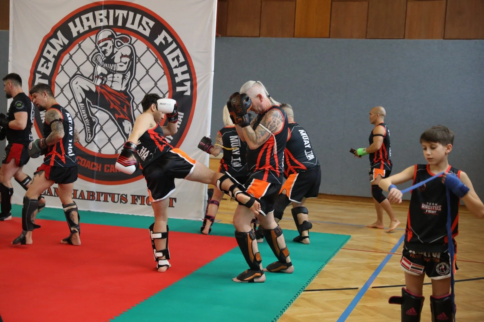 Muay Thai Kampfsporttraining mit Kindern in Fitnesshalle, Trainer und Kinder in Sportkleidung, großes Banner mit Kampfsportlogo im Hintergrund.