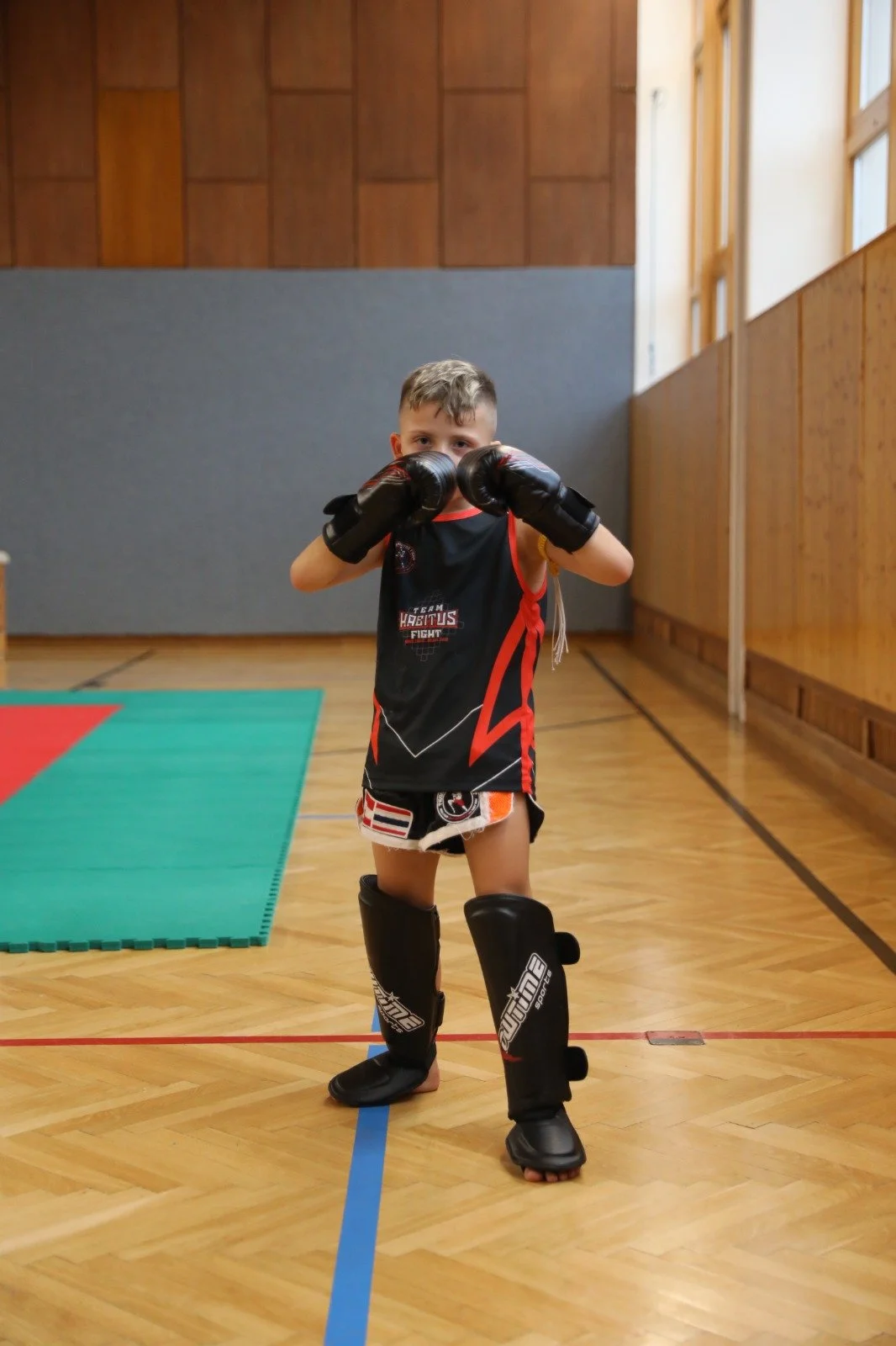 Junge im Muay Thai-Kampfsport-Outfit mit Schienbeinschonern und Boxhandschuhen in einer Turnhalle.