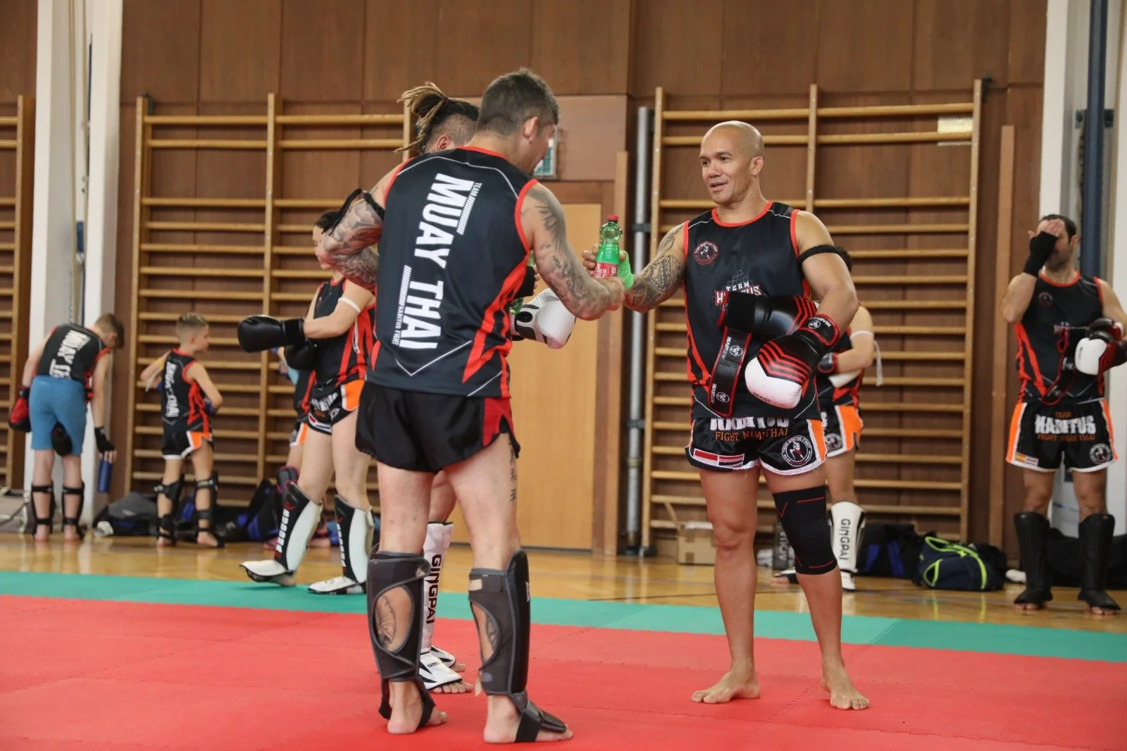 Männer in Muay Thai Kampfausrüstung beim Training in einer Turnhalle, eine Person überreicht einem anderen einen Drink, im Hintergrund Kinder beim Training.