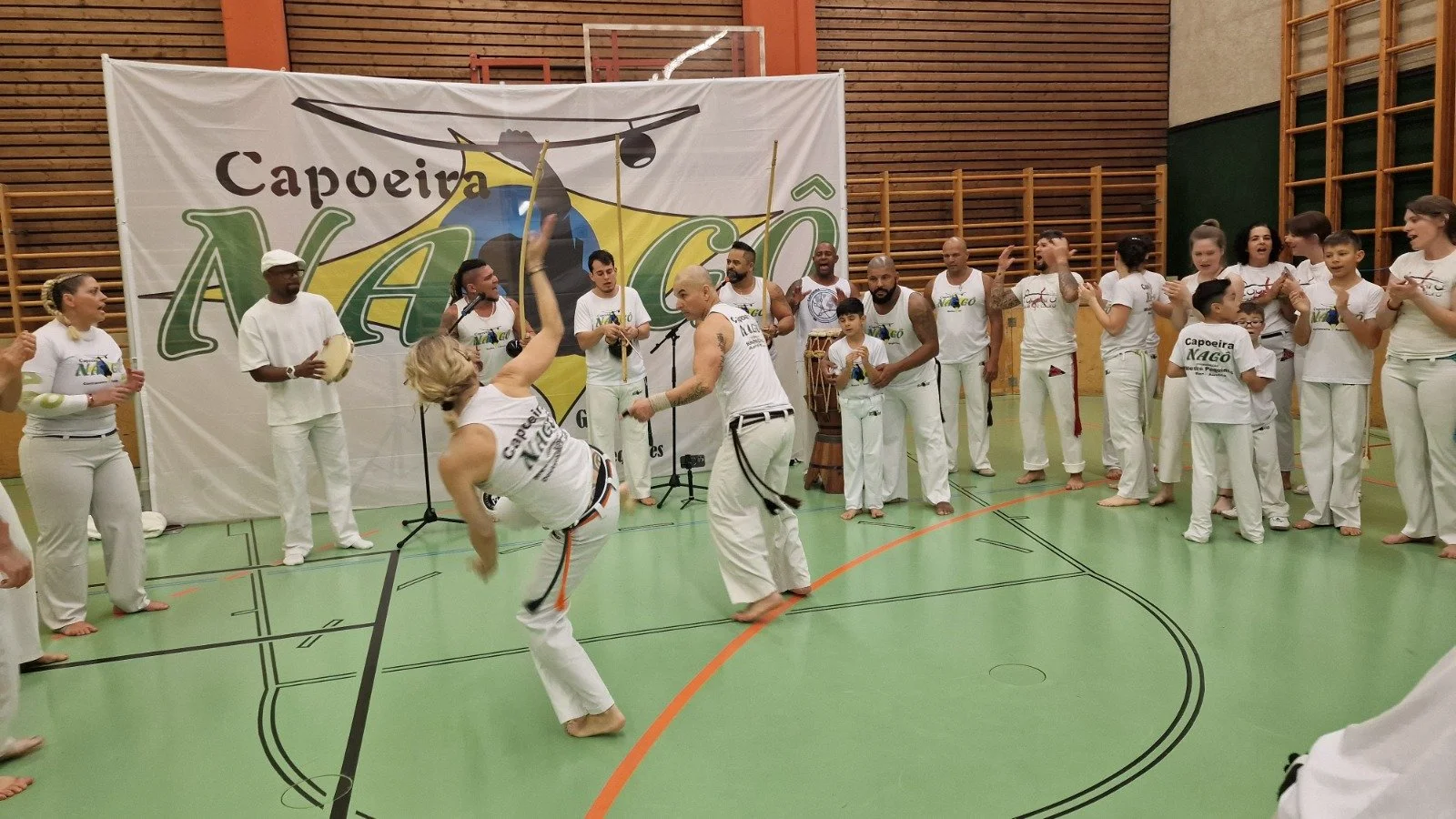 Gruppen von Menschen beim Capoeira in einer Turnhalle, alle in weißer Kleidung, mit einem großen Capoeira NAGO Banner im Hintergrund.