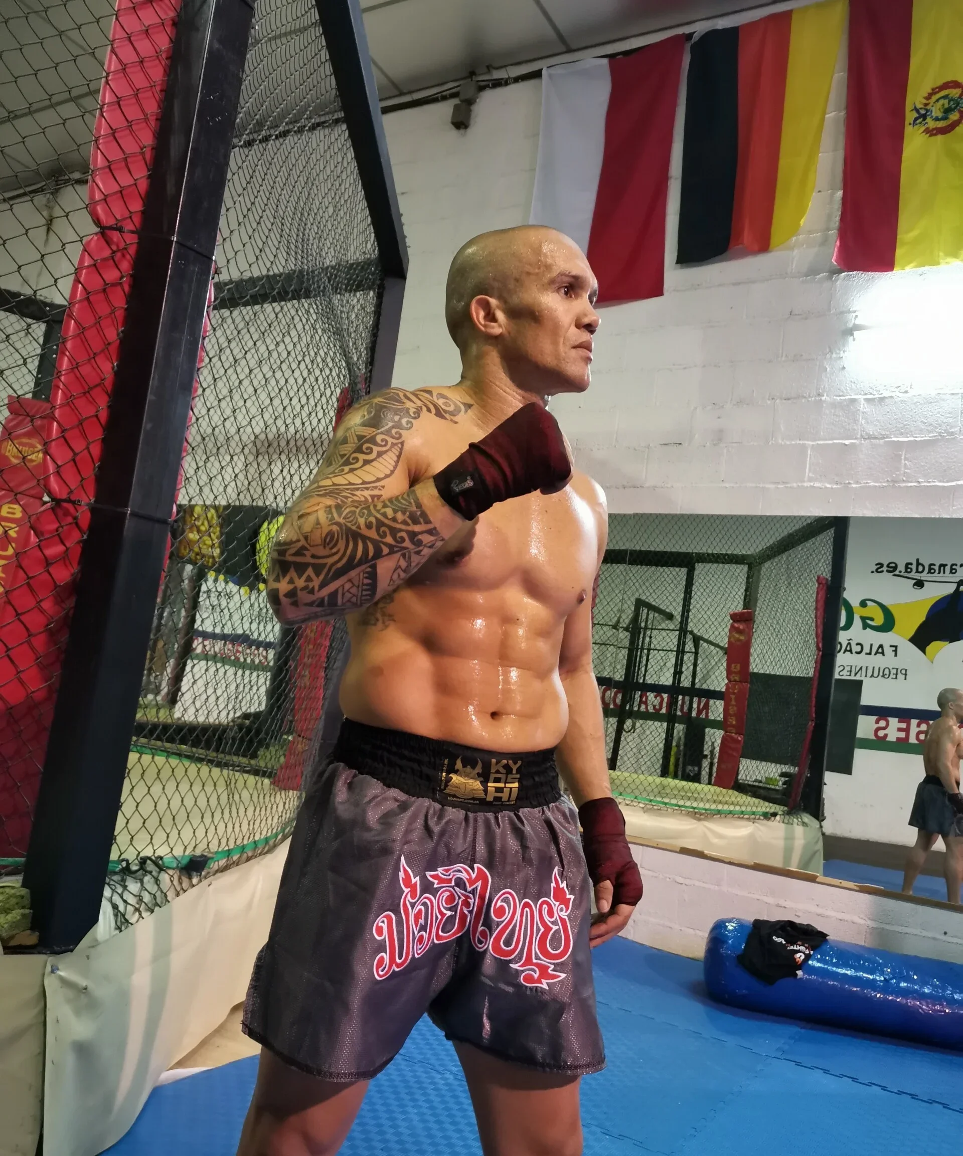 Ein männlicher Boxer in Boxshorts und Handschuhen, mit Tattoos auf den Armen, in einer Trainingshalle.
