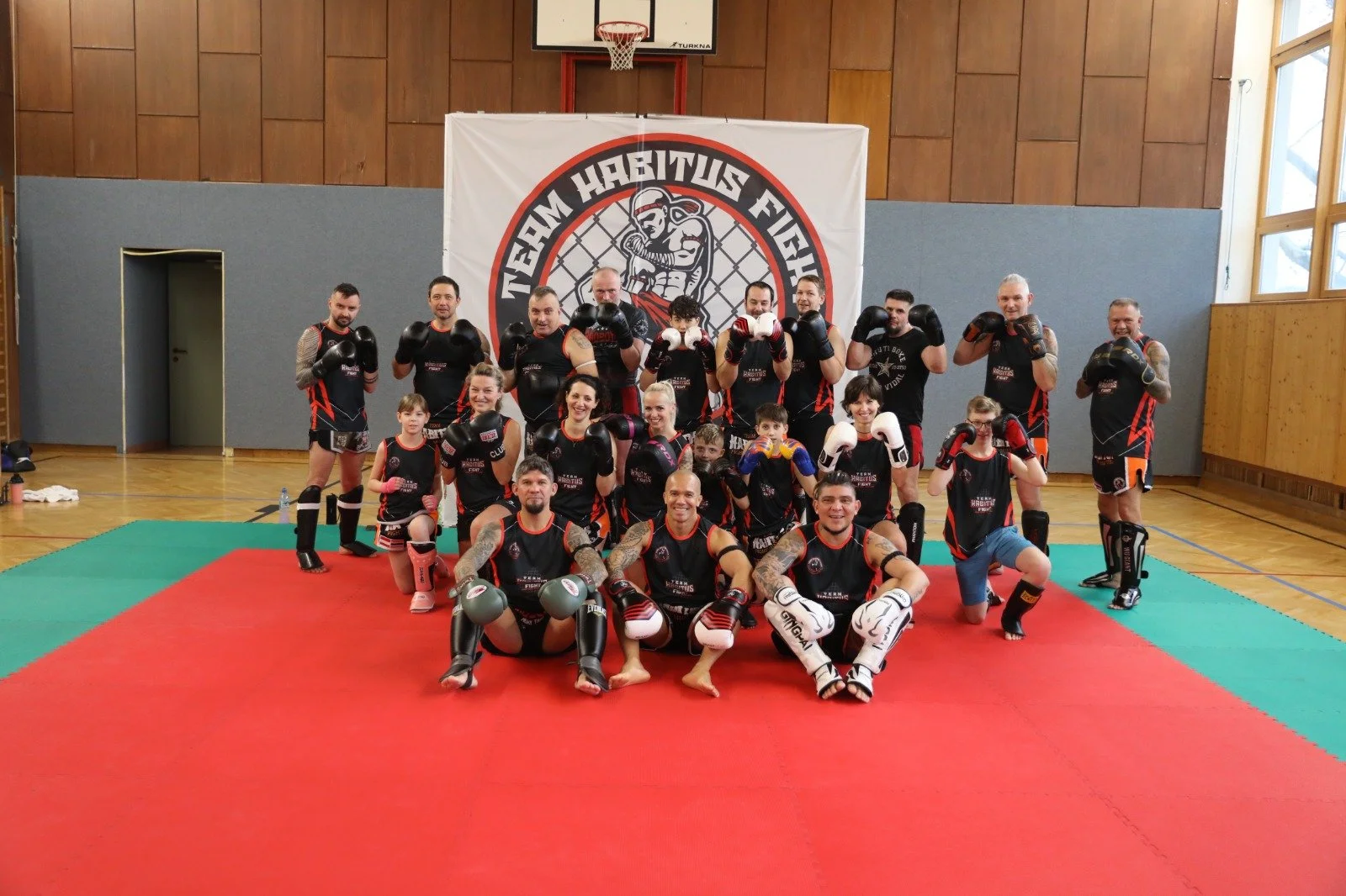 Gruppe von Kampfsportlern und Kindern in Kampfsportbekleidung in einem Sporthalle, die für ein Foto posieren, im Hintergrund ein Banner mit dem Schriftzug "Team Habituus Fight".