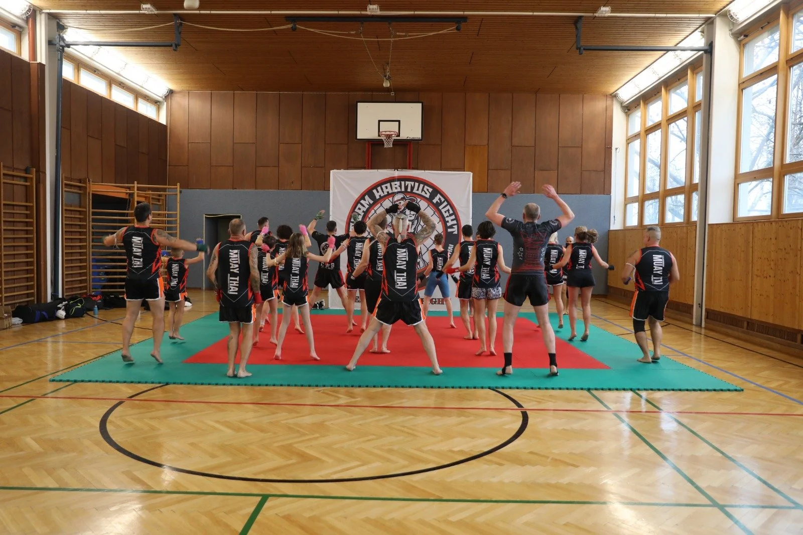 Menschen beim Muay Thai Training in einer Turnhalle, die auf einer roten Turnmatte stehen und sich auf ein Training vorbereiten. Es sind mehrere Teilnehmer in schwarzen Muay Thai-Trikots und ein Trainer dabei.