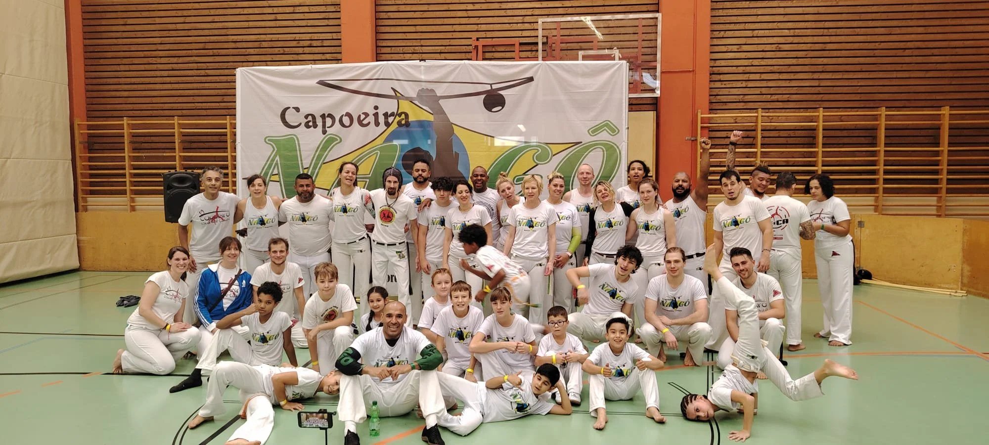 Gruppenfoto von Tae-Kwon-Do und Capoeira Sportlern in einer Turnhalle, alle tragen weiße Sportkleidung mit Logos, einige jubeln mit erhobenen Fäusten, andere sitzen oder liegen am Boden, im Hintergrund ein Banner mit Capoeira und Tae-Kwon-Do Logos.
