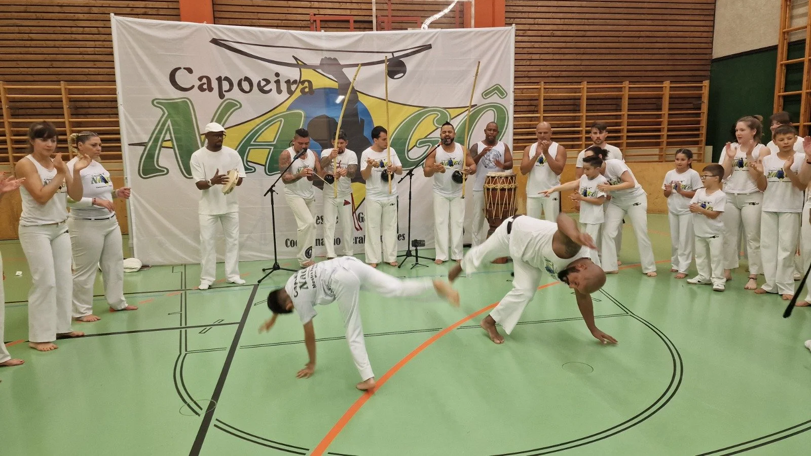 Teilnehmer führen Capoeira-Kampf im Turnhallenraum auf, umgeben von Zuschauern, mit einer großen Banner im Hintergrund, das 'Capoeira NEGO' zeigt.