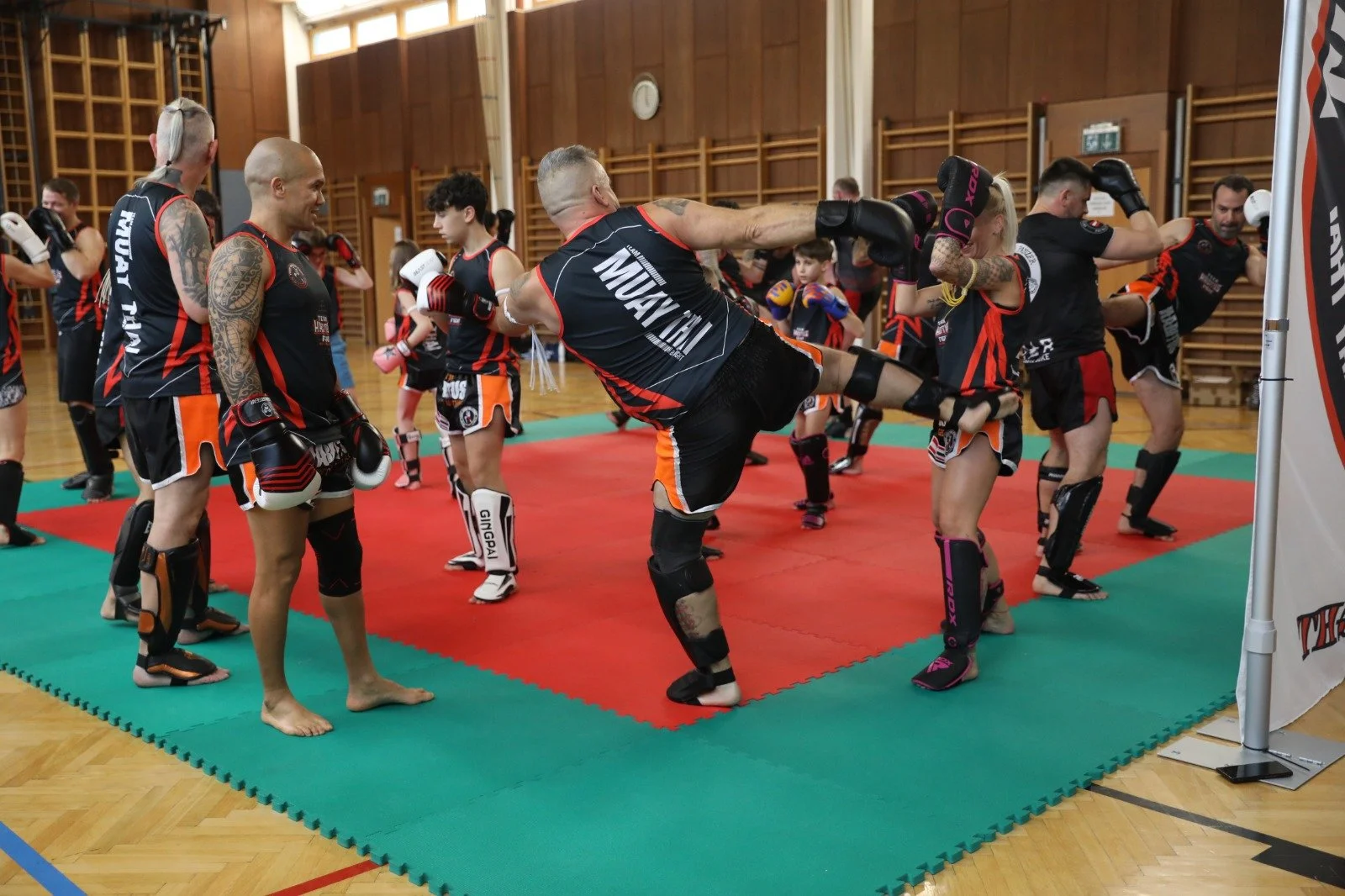 Menschen beim Kickbox-Training in einer Halle, einige üben Schlagtechniken und tragen Schutzausrüstung, darunter Handschuhe, Kopf- und Beinschützer.