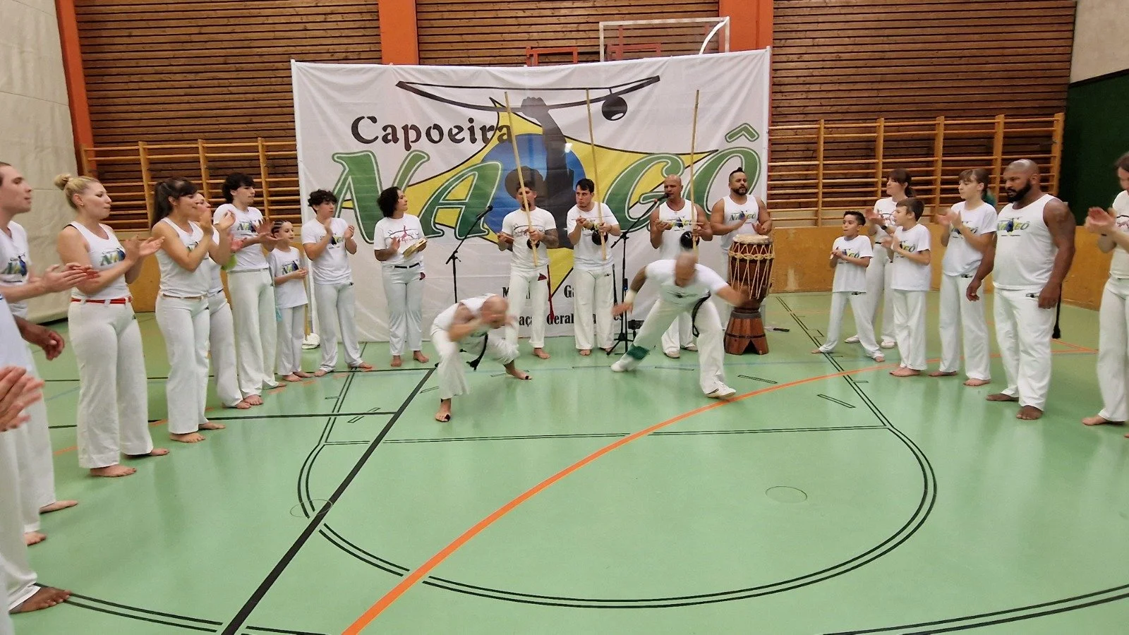 Grupp von Capoeira-Tänzern in einem Turnraum, einige tanzen, andere klatschen oder spielen Instrumente, im Hintergrund ein Banner mit dem Text 'Capoeira'.