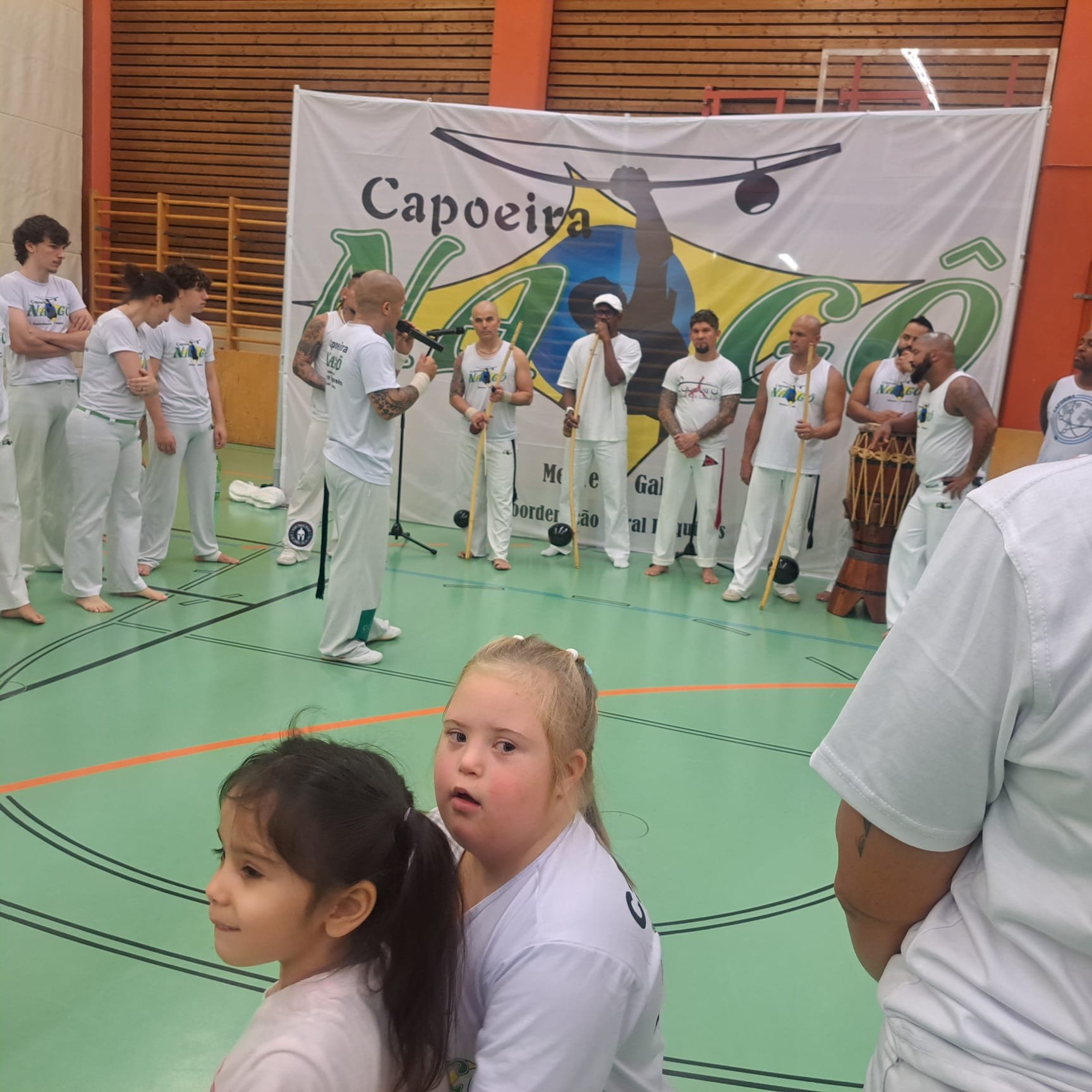 Junge in weißen Kleidung im Capoeira-Workshop, andere Kinder stehen im Hintergrund vor einem großen Capoeira-Banner im Turnhallenzuhause.
