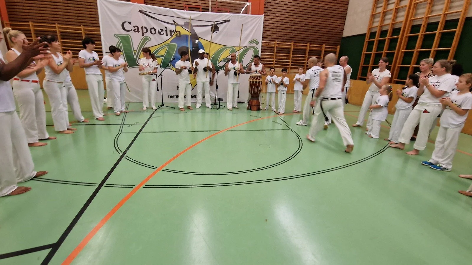 Gruppen von Menschen, vermutlich in einem Sportzentrum, die Capoeira praktizieren oder vorführen. Es gibt Kinder und Erwachsene in weißen Sportkleidung, einige spielen Musikinstrumente, andere klatschen. Ein Banner im Hintergrund trägt den Schriftzug
