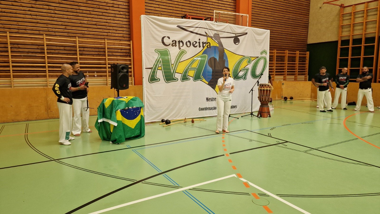 Menschen in einem Turnzaal bei einer Capoeira-Veranstaltung mit einem großen Banner im Hintergrund, das 'Capoeira NAGO' zeigt, und einer Brasilianischen Flagge auf einem Tisch.