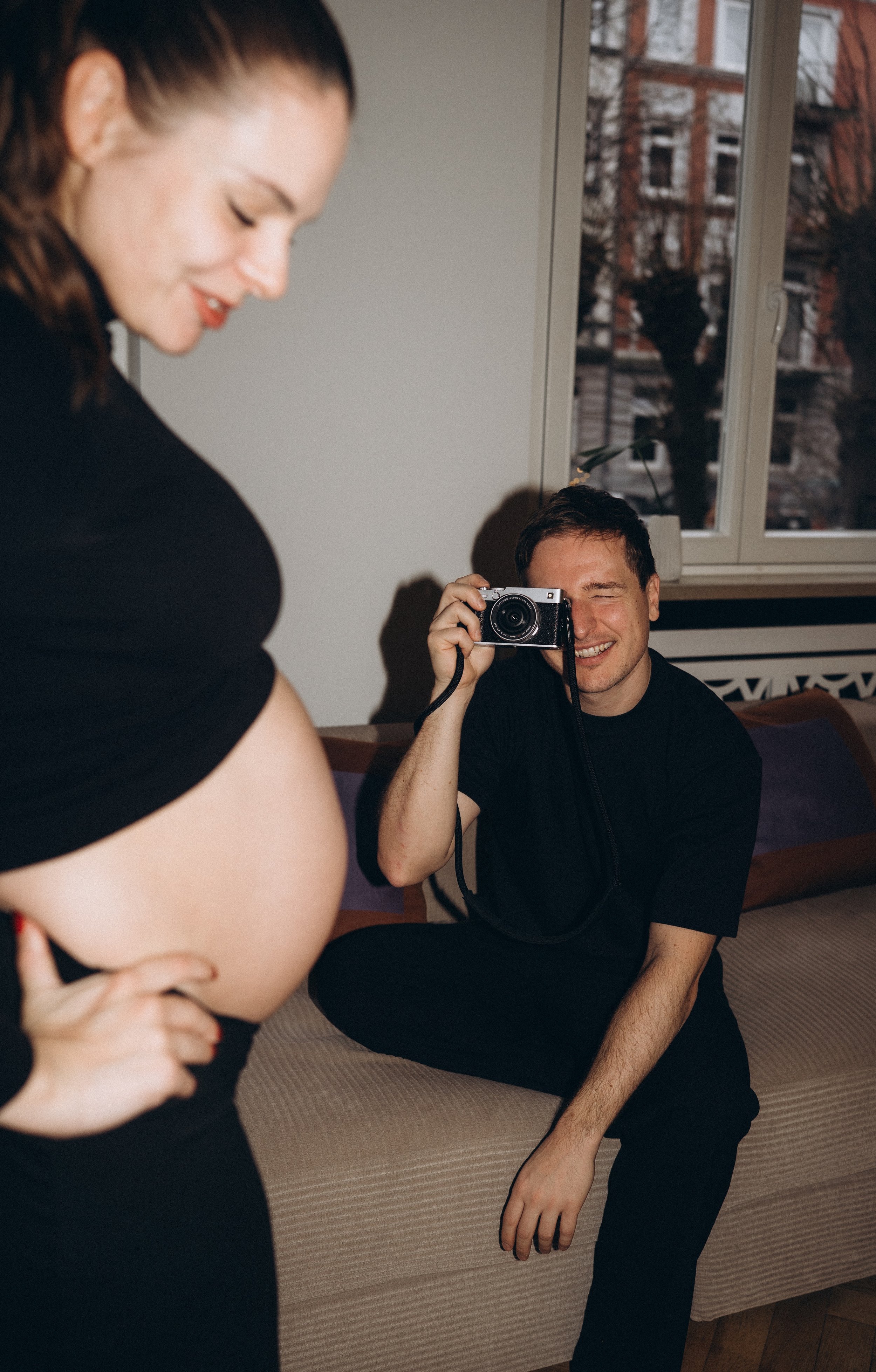 Ein Mann macht ein Foto von einer schwangeren Frau, die ihre Hand auf ihren Bauch legt, in einem Wohnzimmer mit einem Sofa und einem Fenster im Hintergrund.
