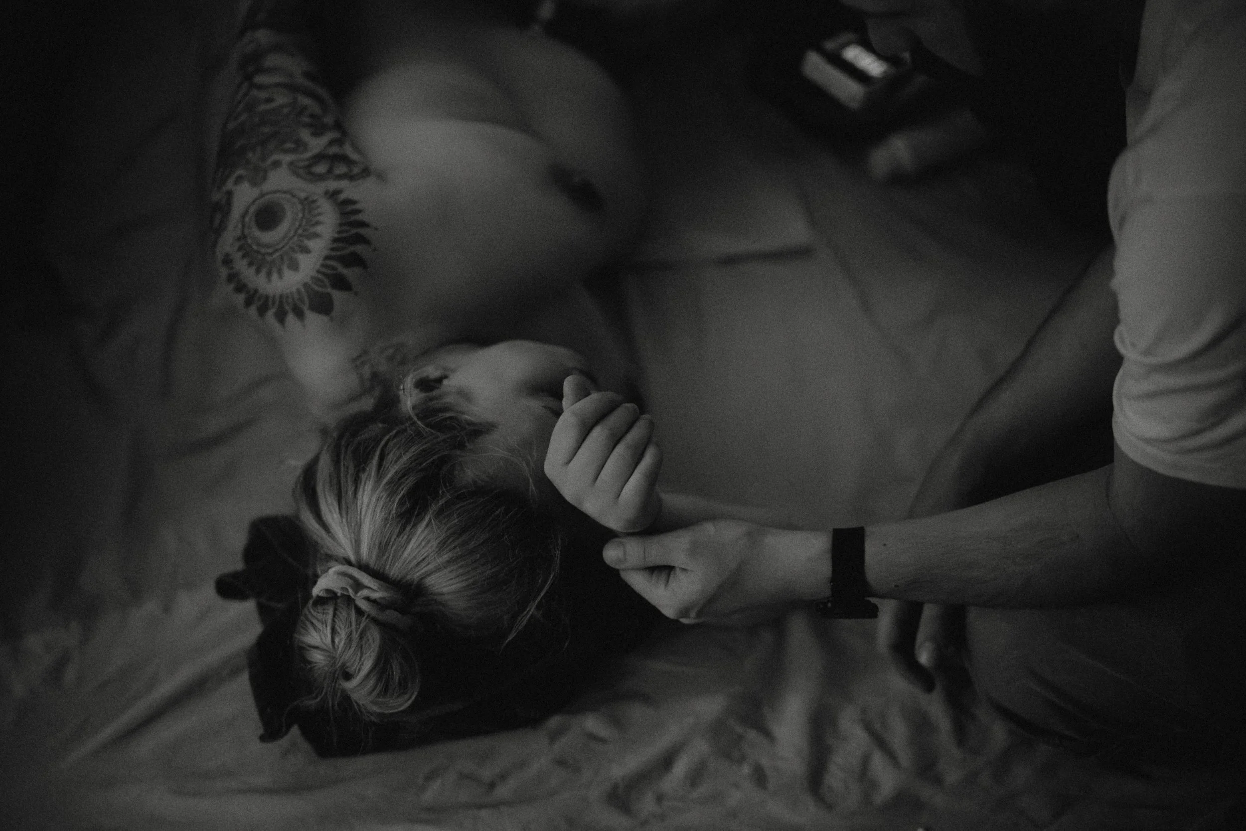 Eine Person liegt auf einem Bett und hält eine andere Person an der Hand, während die zweite Person auf dem Rücken liegt. Das Bild ist in Schwarzweiß.