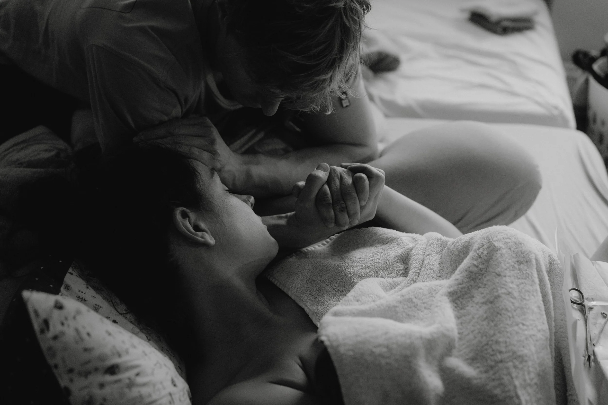 Eine Frau liegt im Krankenbett mit einem Tuch um die Schultern, während ein Mann neben ihr sitzt und ihre Hand hält, in einem schwarz-weißen Foto.