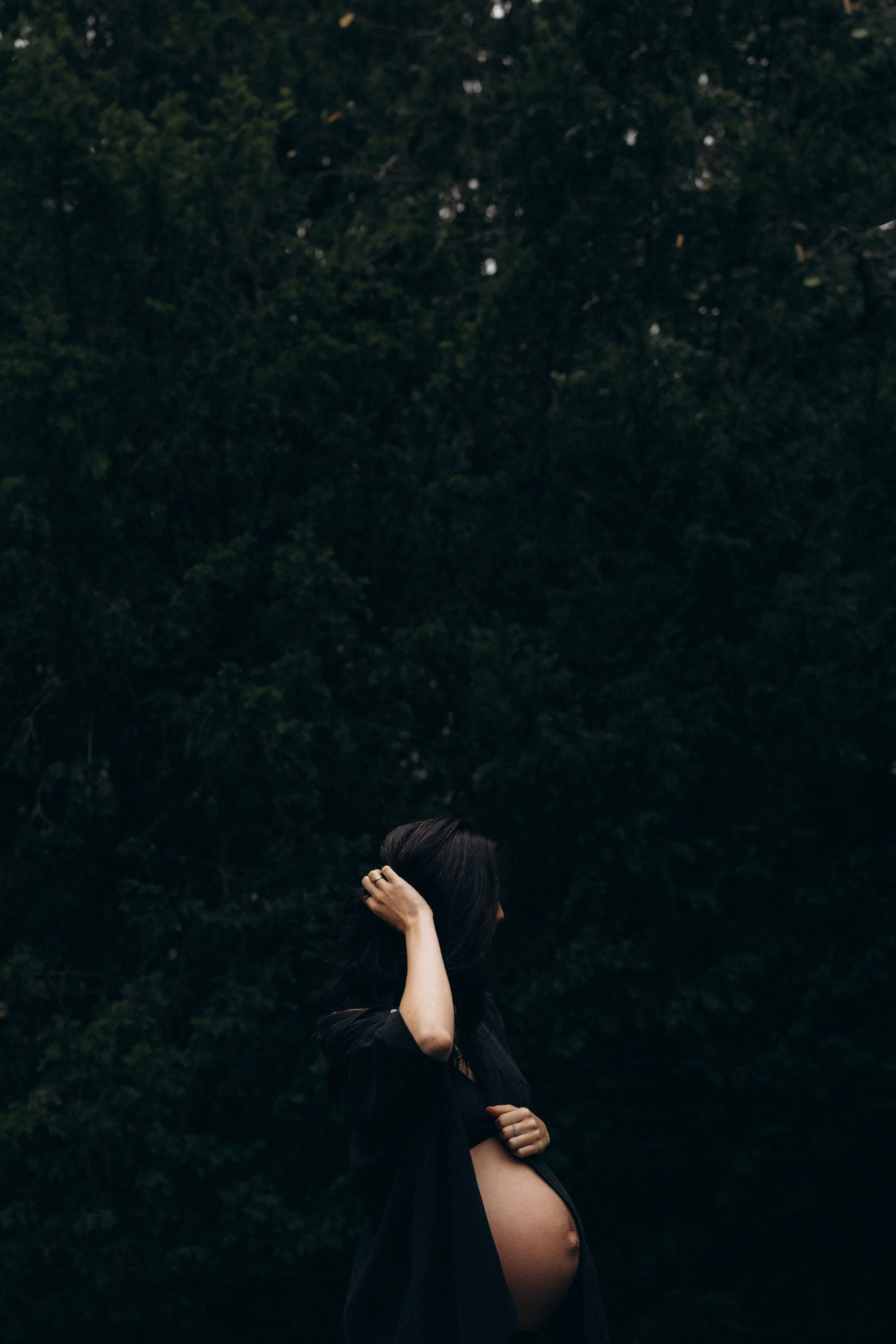 Schwangere Frau steht in einem dunklen Wald, trägt eine schwarze Jacke, die sie teilweise offen trägt, und berührt ihren Bauch mit einer Hand, während sie die andere Hand im Haar hält.
