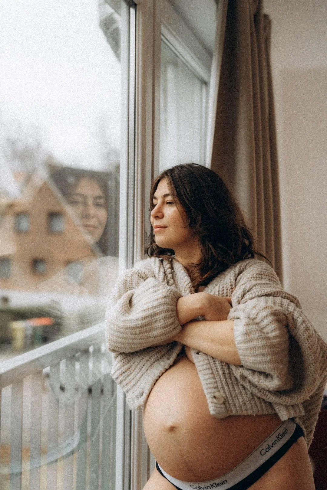 Schwangere Frau schaut nach draußen durch ein Fenster, trägt bequeminierende Kleidung, inklusive Calvin Klein Unterhose, und lehnt sich entspannt gegen das Fenster.