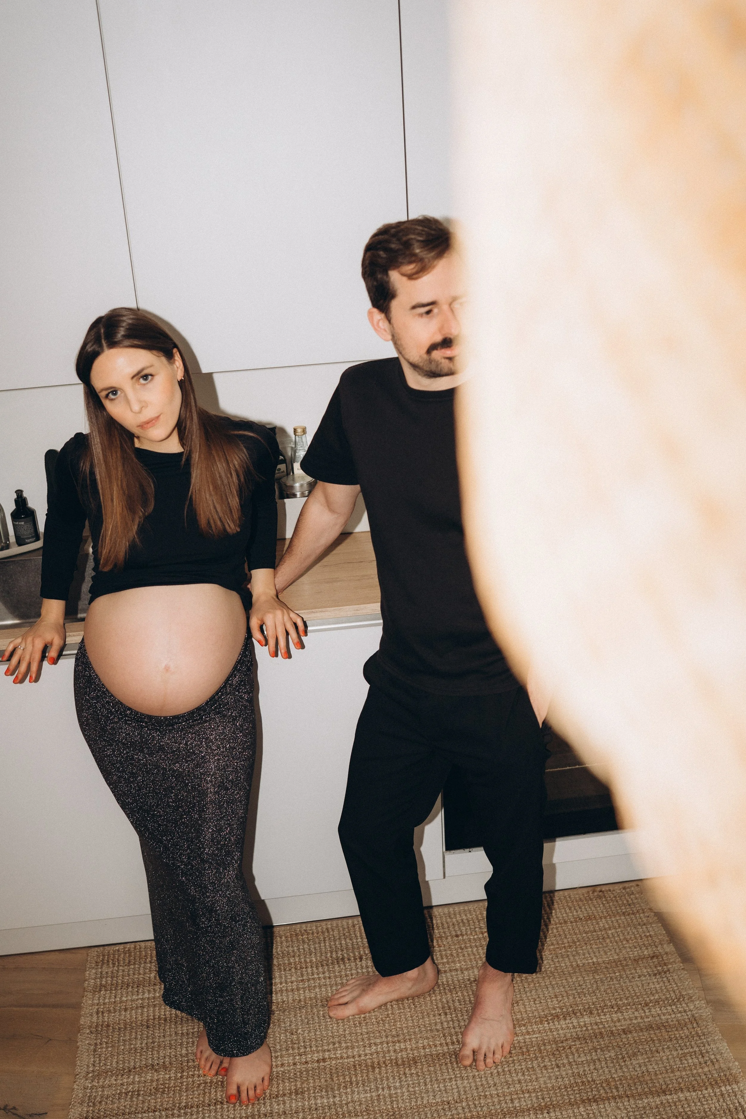 Eine schwangere Frau lehnt sich an die Küchenarbeitsplatte, während ein Mann neben ihr steht, beide in einer modernen Küche, die Szene ist humorvoll und leger.