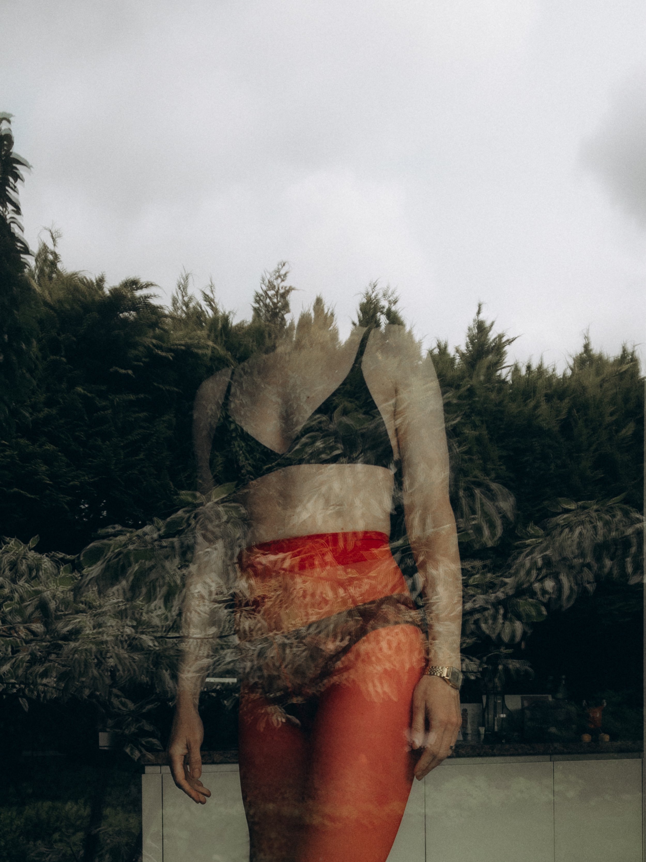 Reflexion einer Frau im Fenster mit schwarzem Sport-BH und roten Leggings, umgeben von Bäumen, an einem bewölkten Tag.