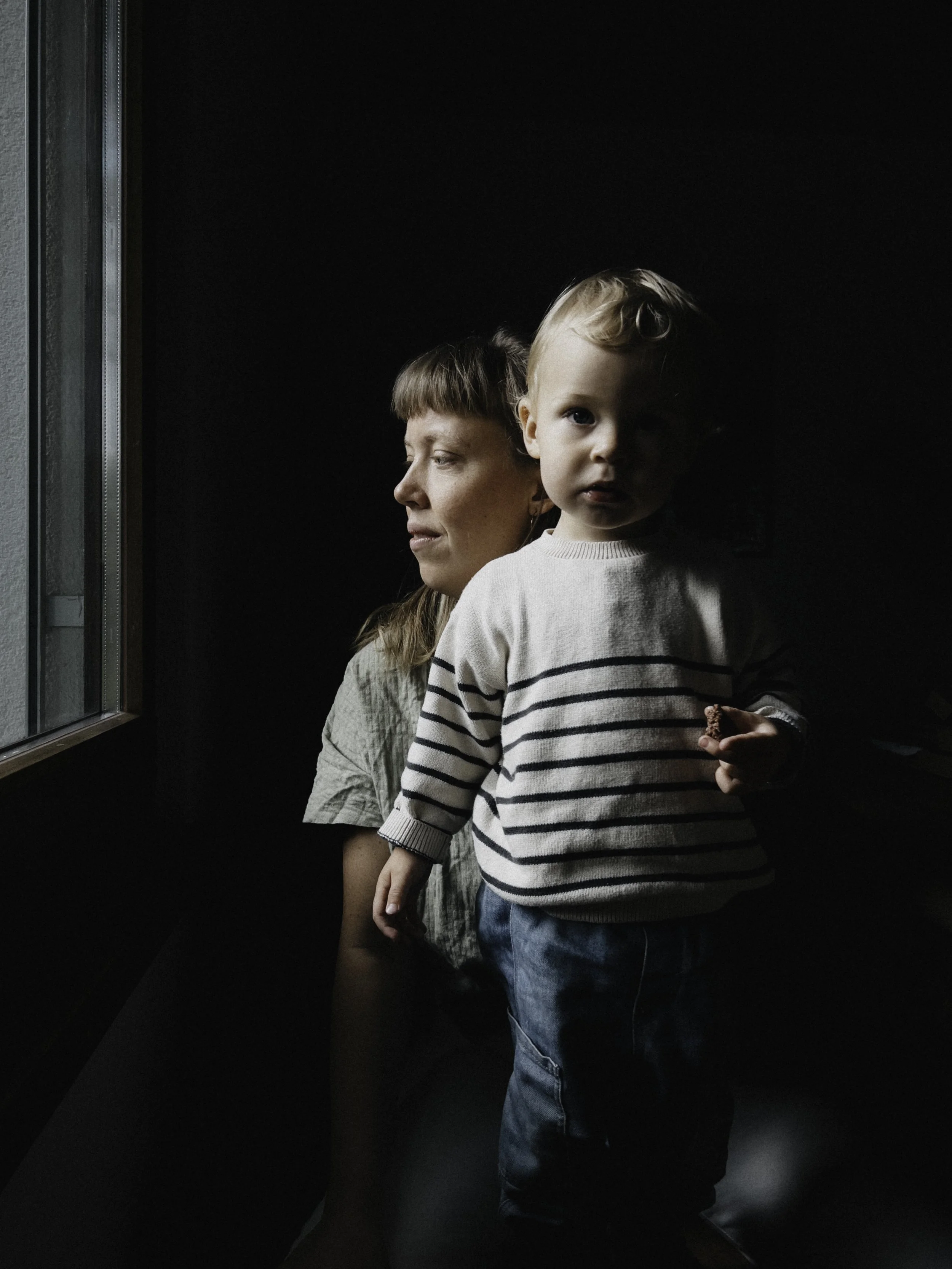 Eine Frau und ein kleines Kind stehen vor einem Fenster in einem dunklen Raum, die Frau schaut nach draußen, während das Kind direkt in die Kamera schaut.