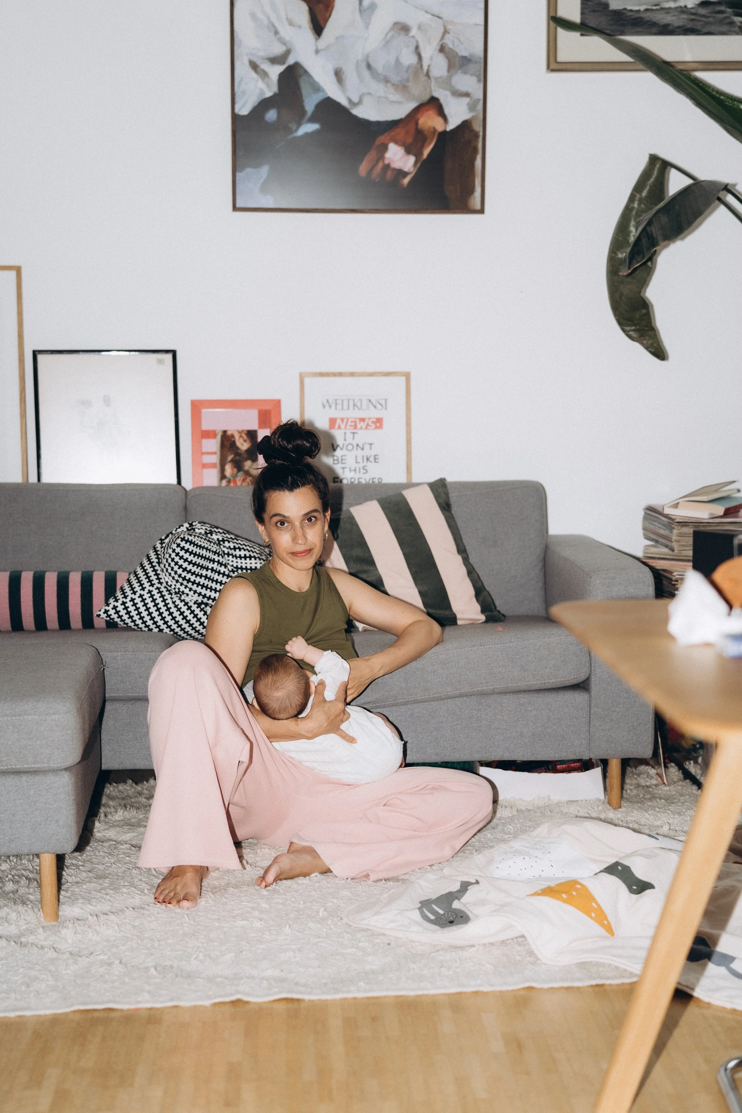 Eine Frau sitzt auf dem Boden vor einem grauen Sofa und stillt ein Baby, während sie im Wohnzimmer entspannt aussieht.