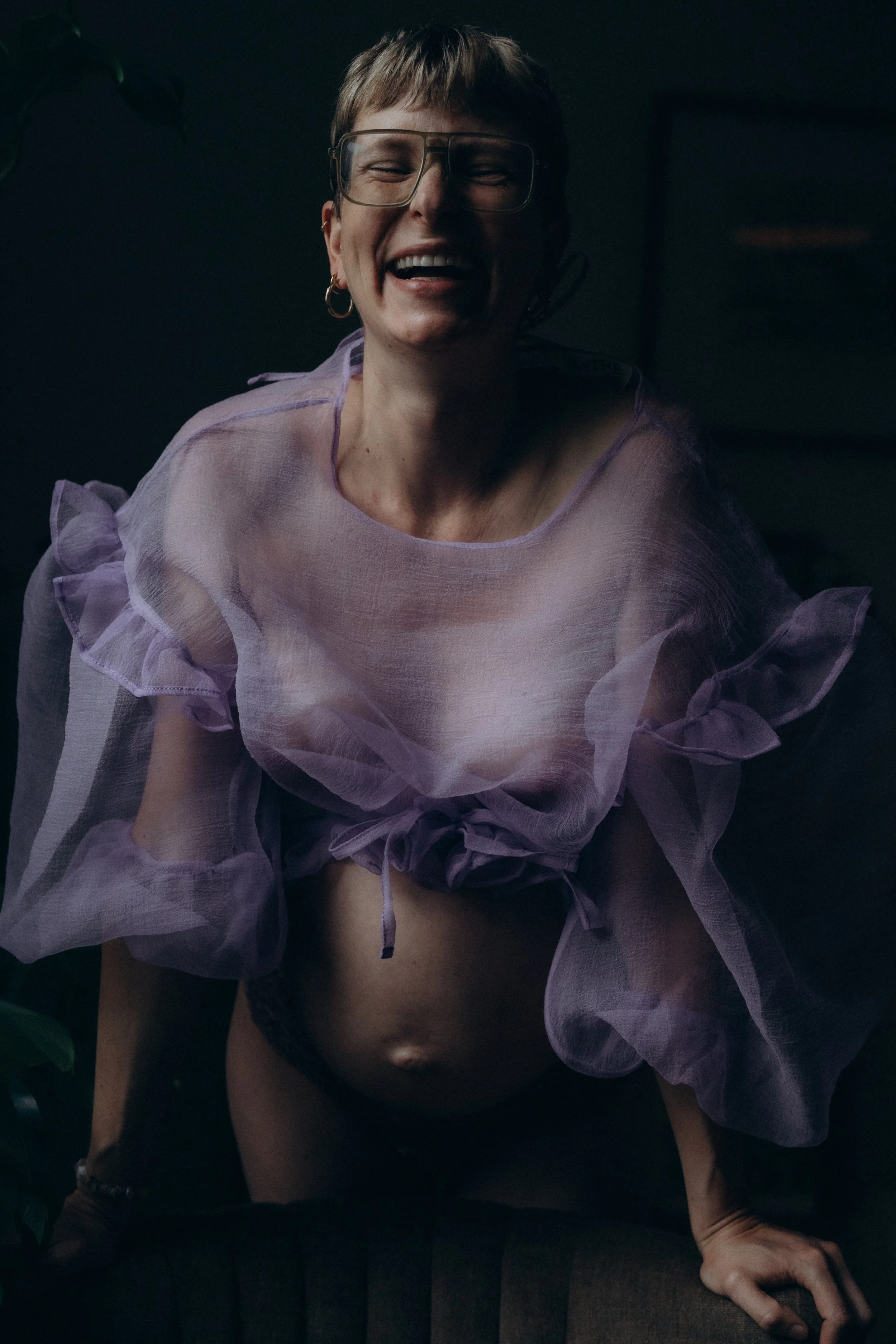 Lachende Frau mit Brille und kurzem Haar, trägt einen lilafarbenen, transparenten blouse, die ihren Bauch freilegt, sitzt auf einem Stuhl in einem dunklen Raum.