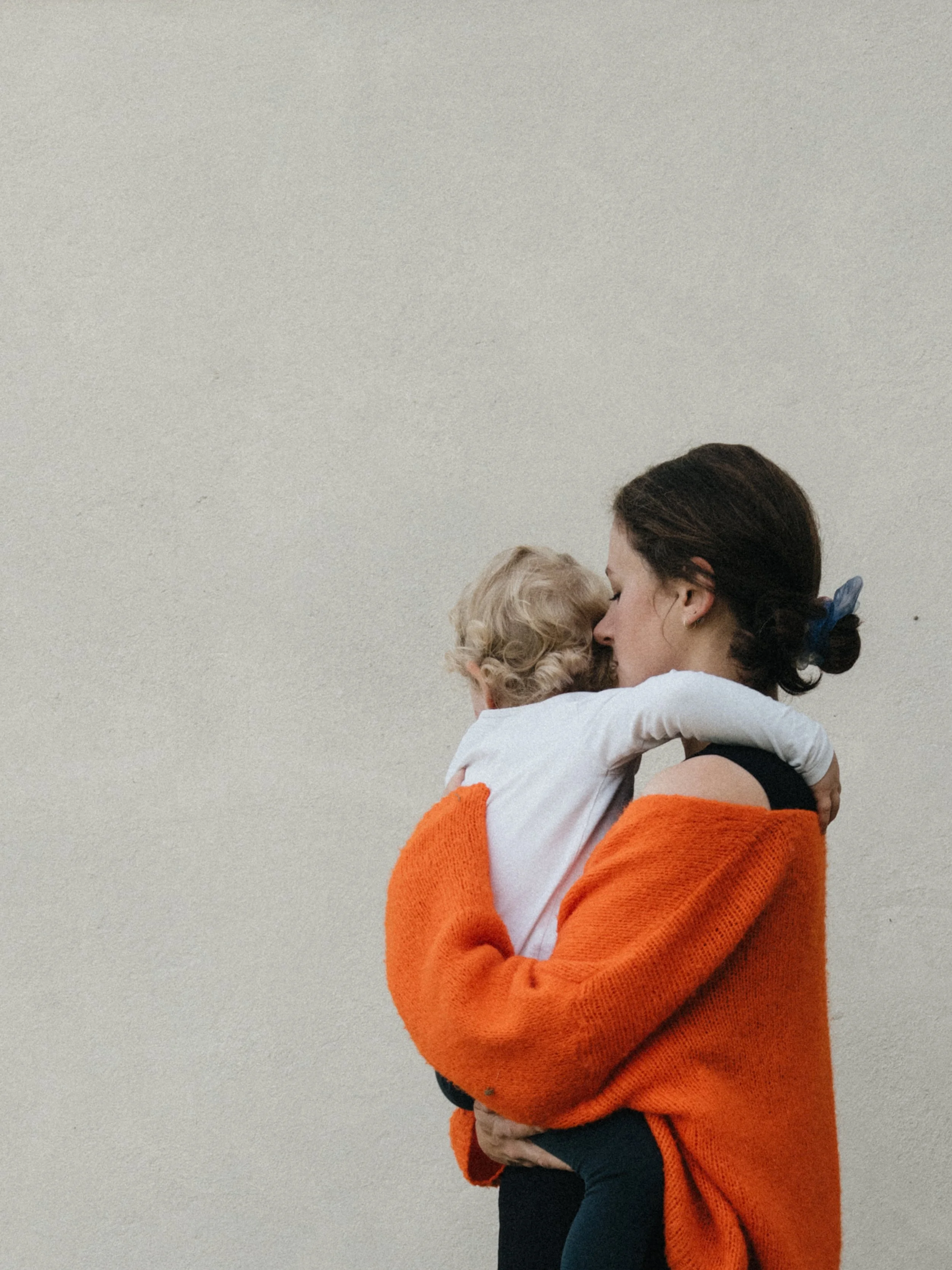 Eine Frau trägt ein kleines Kind, sie umarmen sich liebevoll vor einer neutralen Wand.