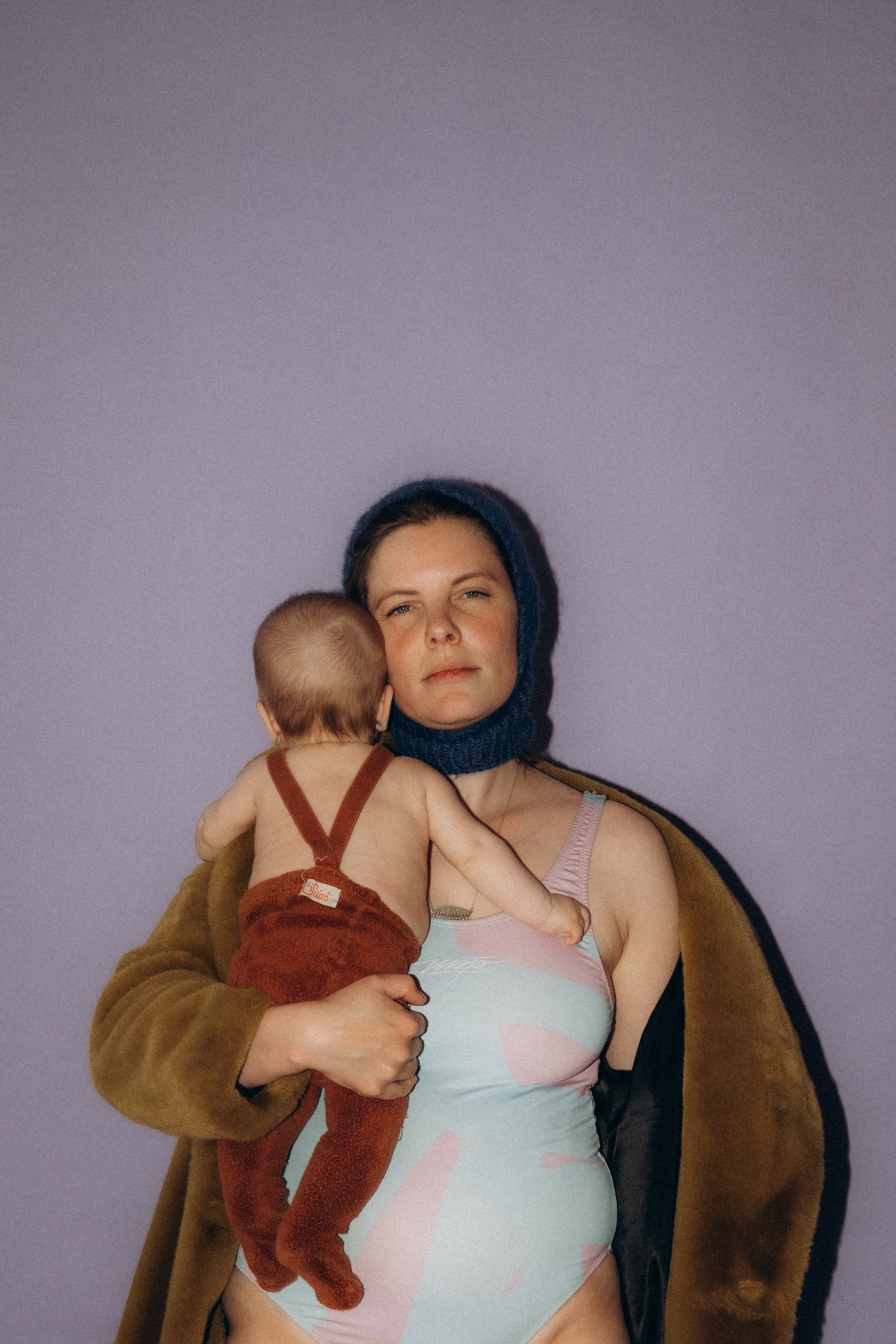 Eine Frau hält ein Baby, das den Rücken zur Kamera hat. Die Frau trägt einen Haarschal und ein Bustier, während das Baby keine Oberbekleidung trägt und ein rotes Kleid trägt.