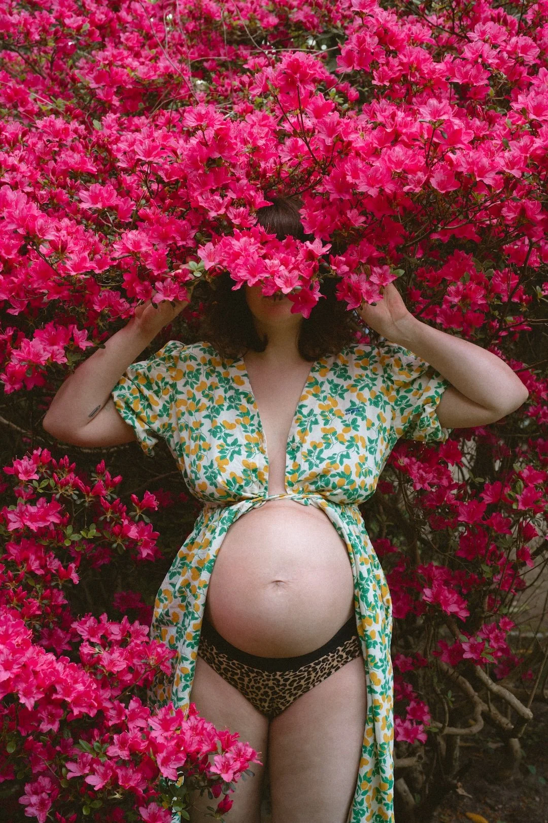 Frau mit Babybauch steht vor einer großen rosa Blütenbüschel, verhüllt das Gesicht mit den Blumen, trägt ein vorgebundenes Kleid mit grünen und gelben Mustern und Leoparden-Print-Unterwäsche.