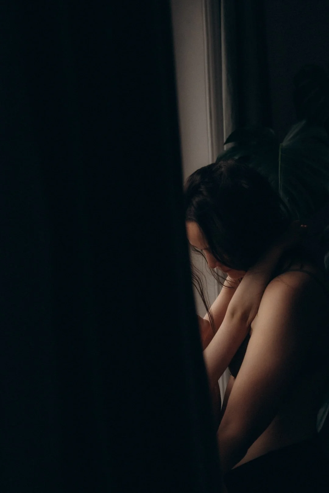 Eine junge Frau mit dunklen Haaren sitzt im Dunkeln, den Kopf auf den Armen auf einem Boden oder Sofa, wobei sie nach unten schaut.