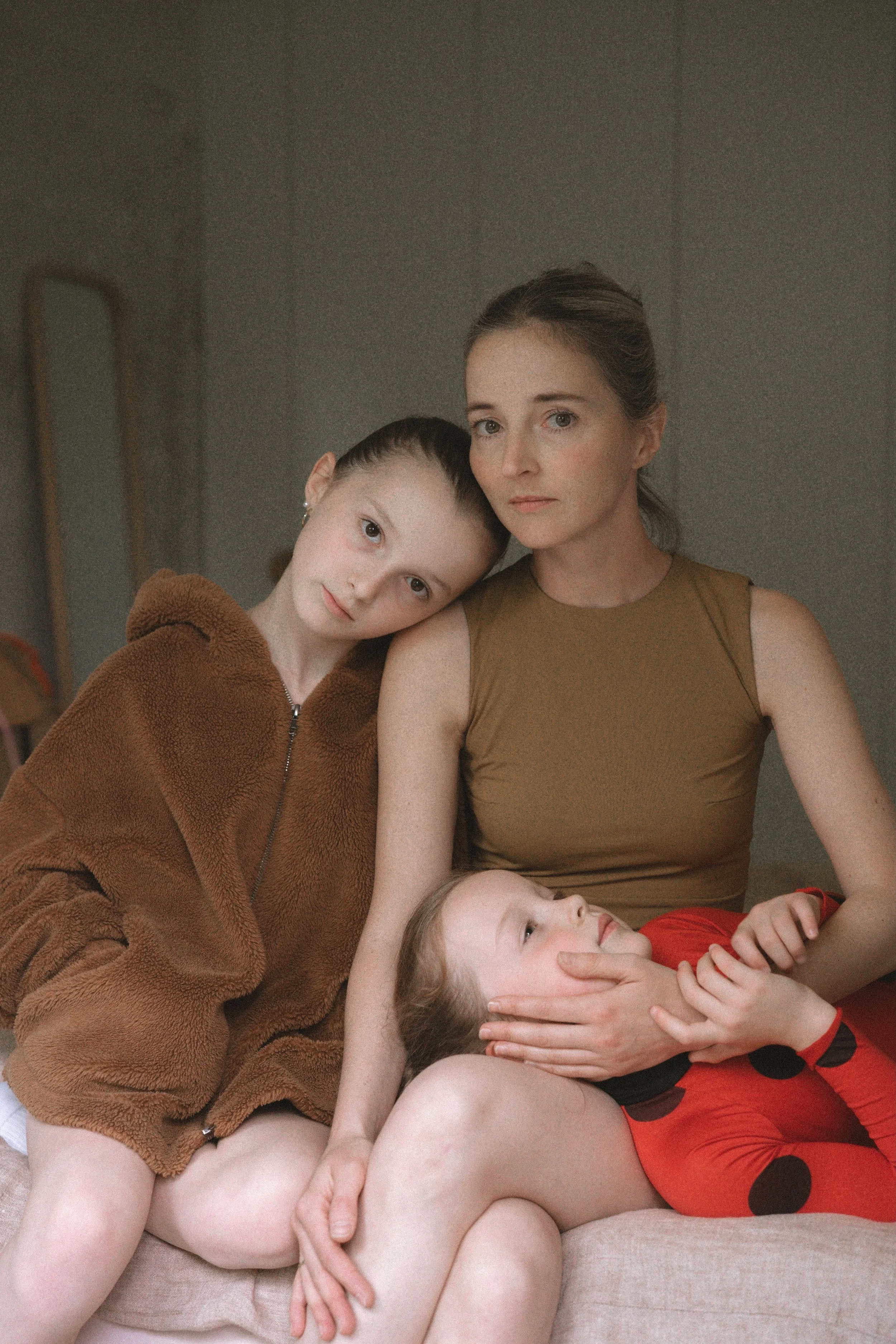 Eine Frau sitzt mit zwei Kindern auf einem Sofa. Das Mädchen auf der linken Seite trägt eine braune Fleecejacke, die andere sitzt auf den Knien der Frau und trägt rote Kleidung mit schwarzen Punkten.