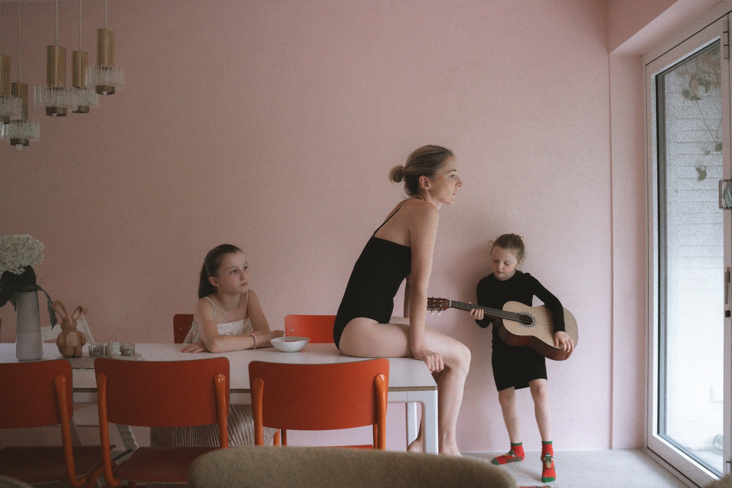Frau in schwarzem Body sitzt auf einem Esstisch, während zwei Kinder mit Gitarre in einem Raum mit rosa Wand spielen und zuhören.