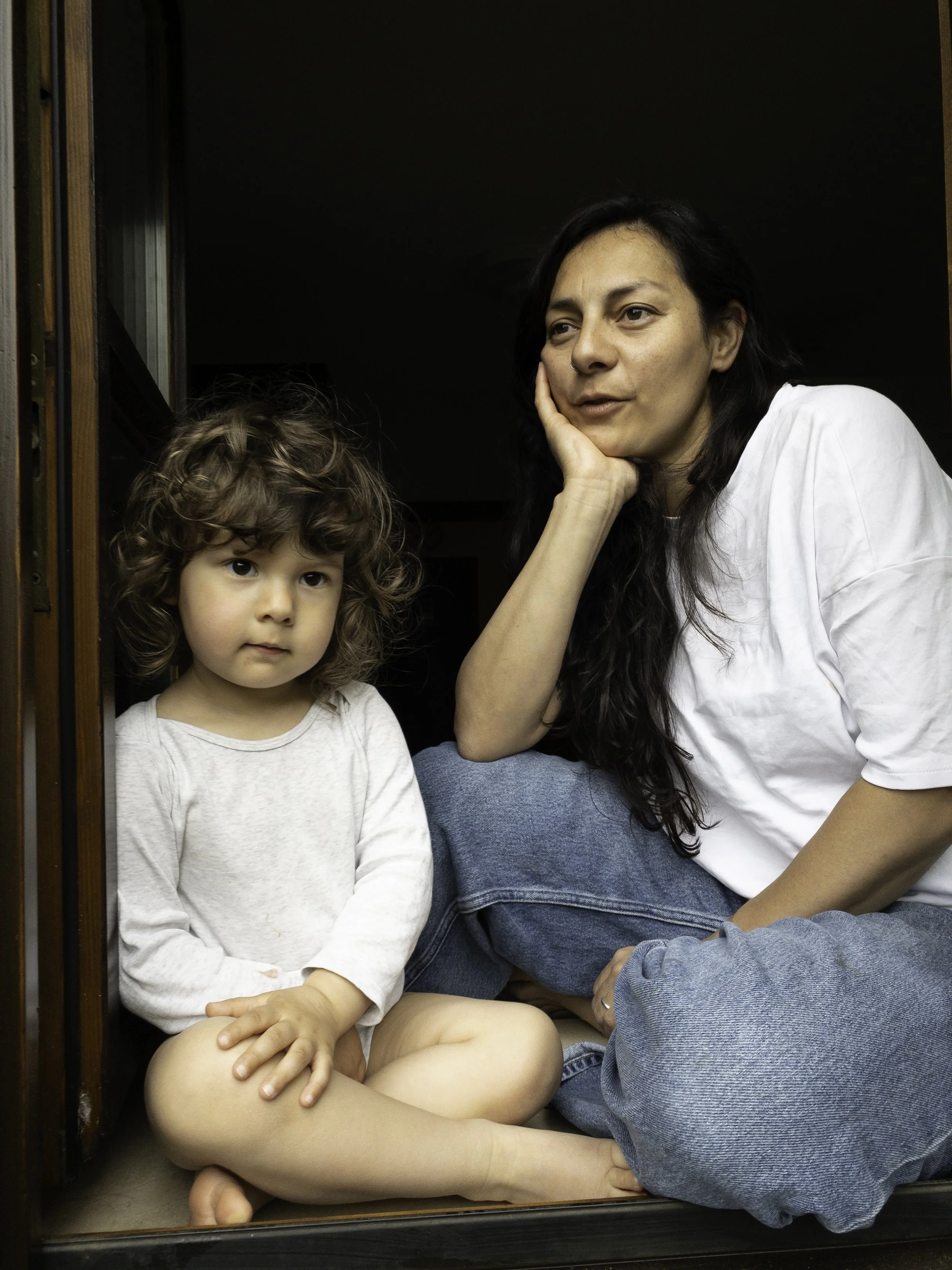 Eine Frau sitzt auf dem Boden mit einem Kind, beide schauen nach vorne, sitzen im dunklen Raum vor einer dunklen Wand, die Frau trägt ein weißes T-Shirt, das Kind trägt ein langärmeliges weißes Shirt, das Kind hat lockige braune Haare.