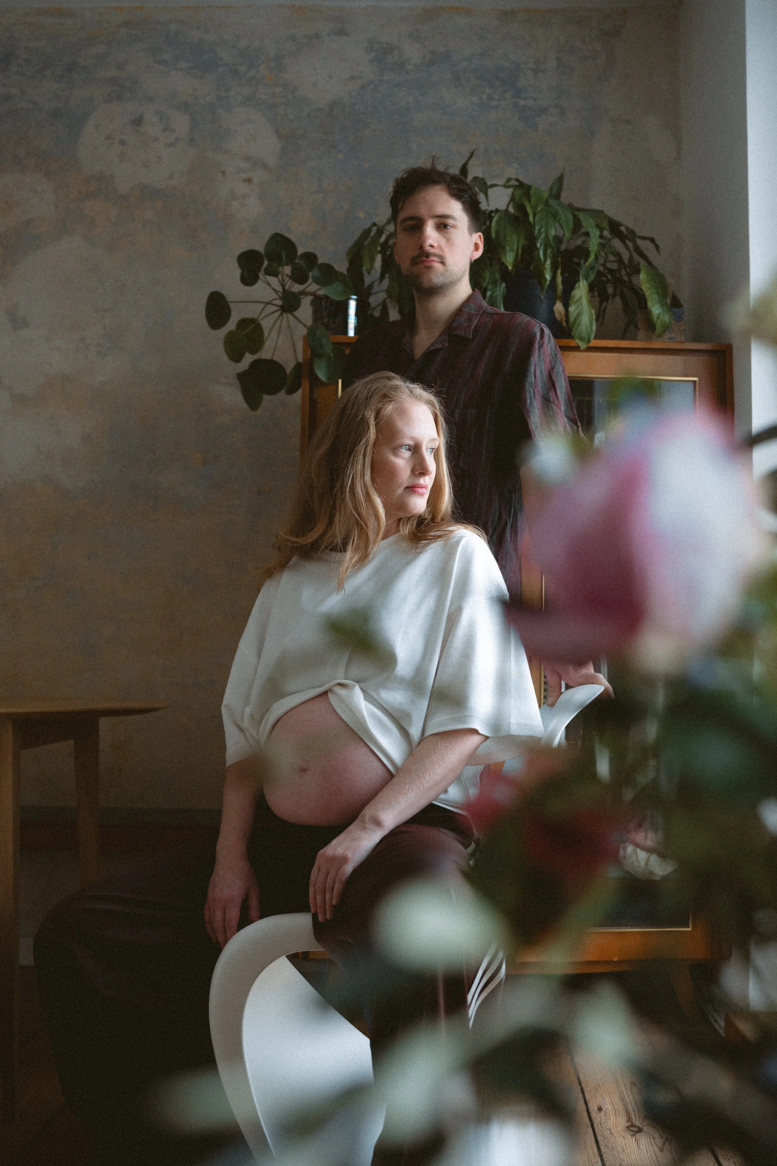 Eine schwangere Frau mit langem, lockigem, rotem Haar sitzt auf einem weißen Stuhl und schaut nach rechts. Ein Mann mit dunklen, kurzen Haaren steht hinter ihr, blickt direkt in die Kamera. Im Vordergrund sind unscharfe Blumen, die die Szene kunstvol