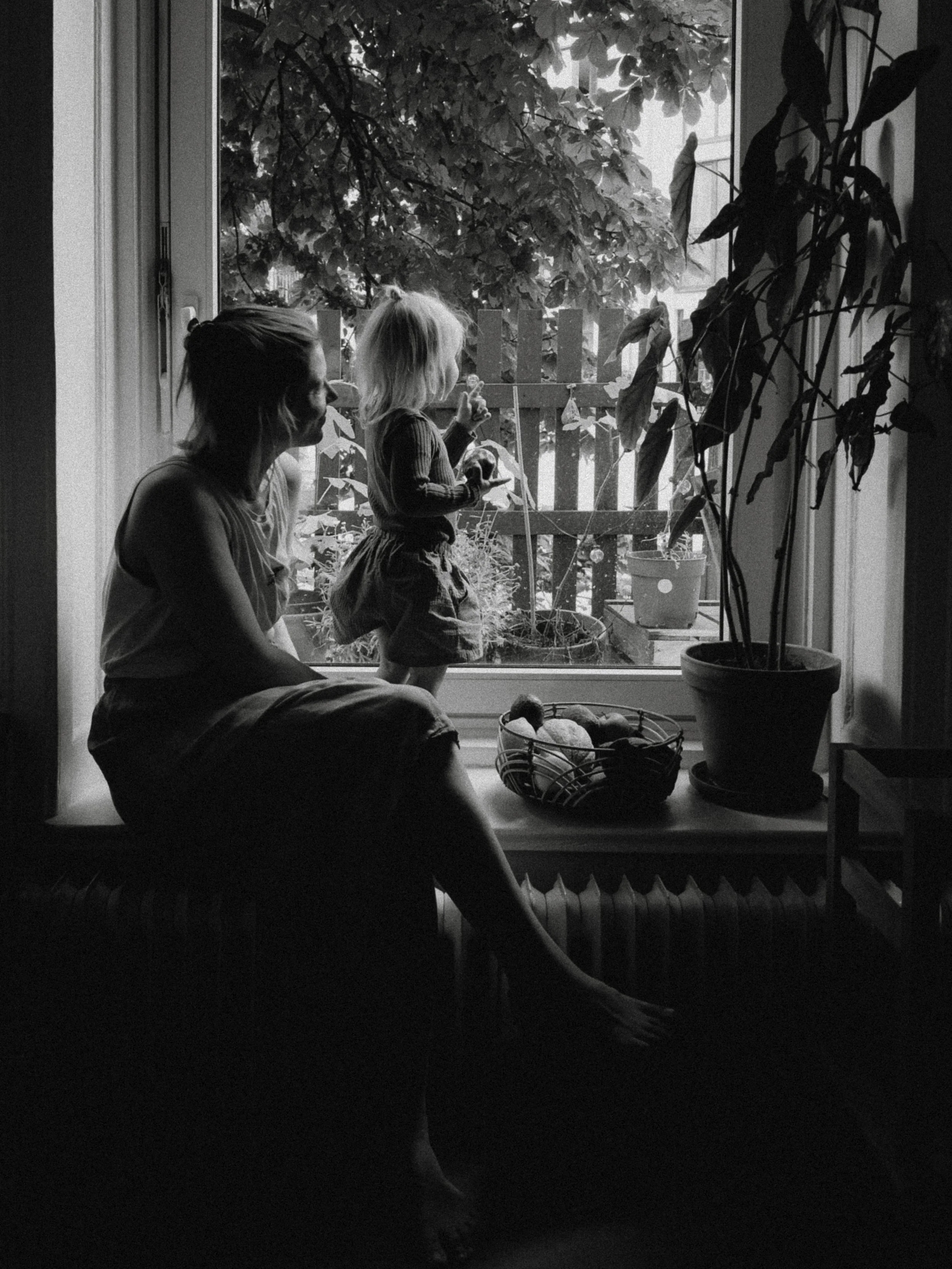 Eine Frau sitzt auf einem Fensterbank, während ein kleines Mädchen auf dem Fensterrahmen steht und nach draußen schaut, in einem schwarz-weißen Bild.