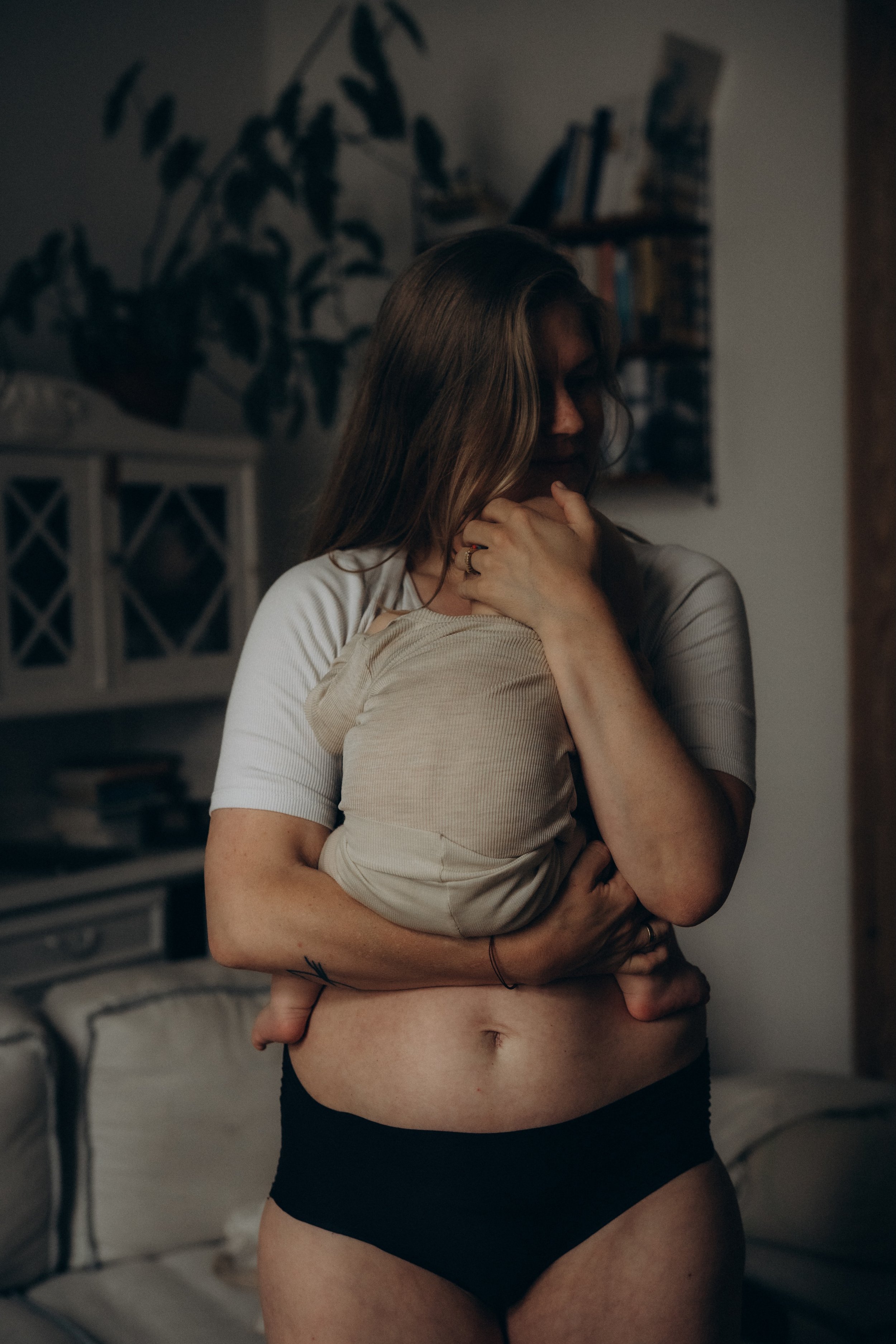 Eine Frau hält ein Baby im Arm in einem Wohnzimmer, both in einem dunklen, warmen Licht, ohne Schuhe, im Hintergrund Bücherregale und Pflanzen.