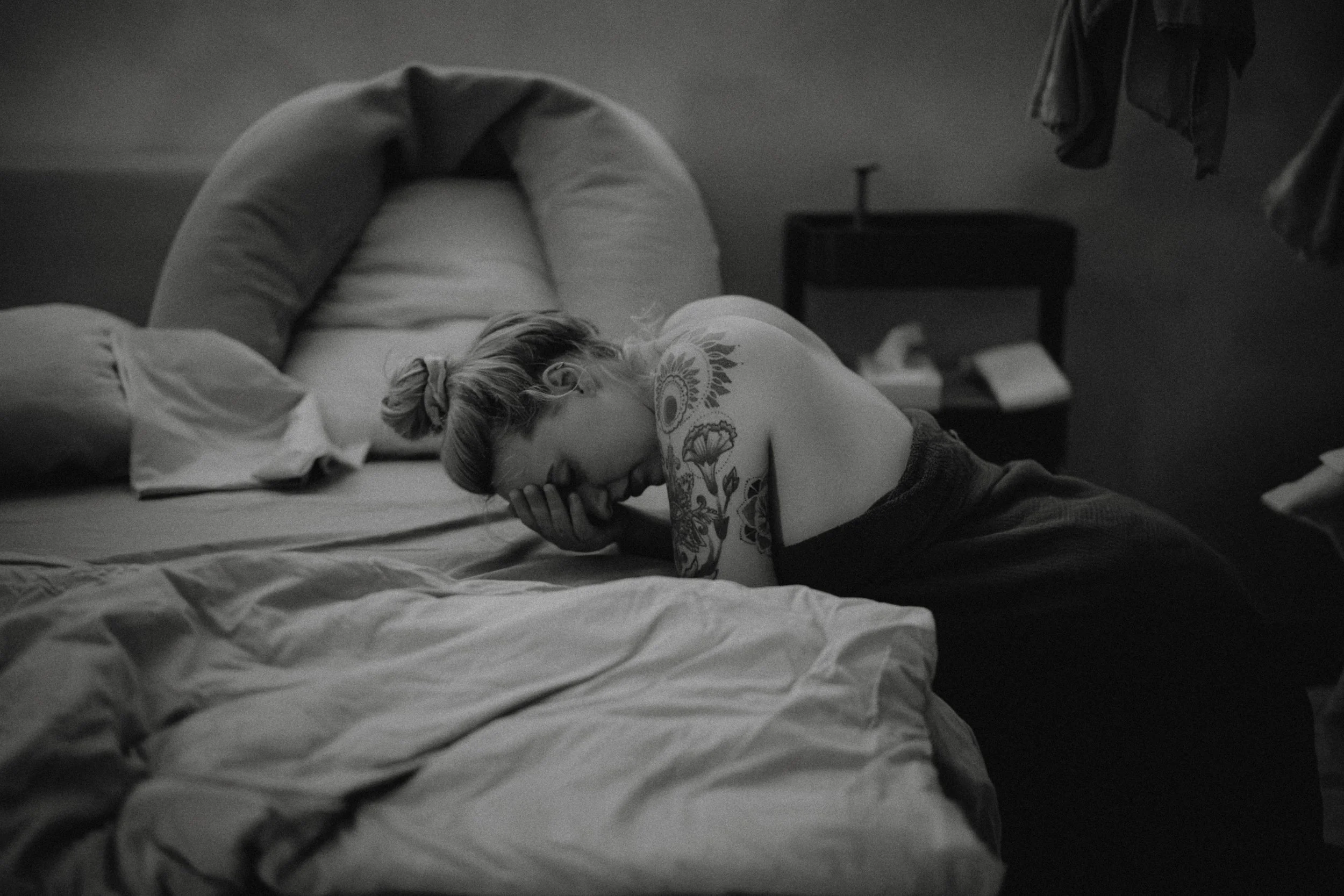 Eine Frau mit Tattoo auf dem Arm liegt auf einem Bett, den Kopf in die Hand gestützt, in einem dunklen Raum.