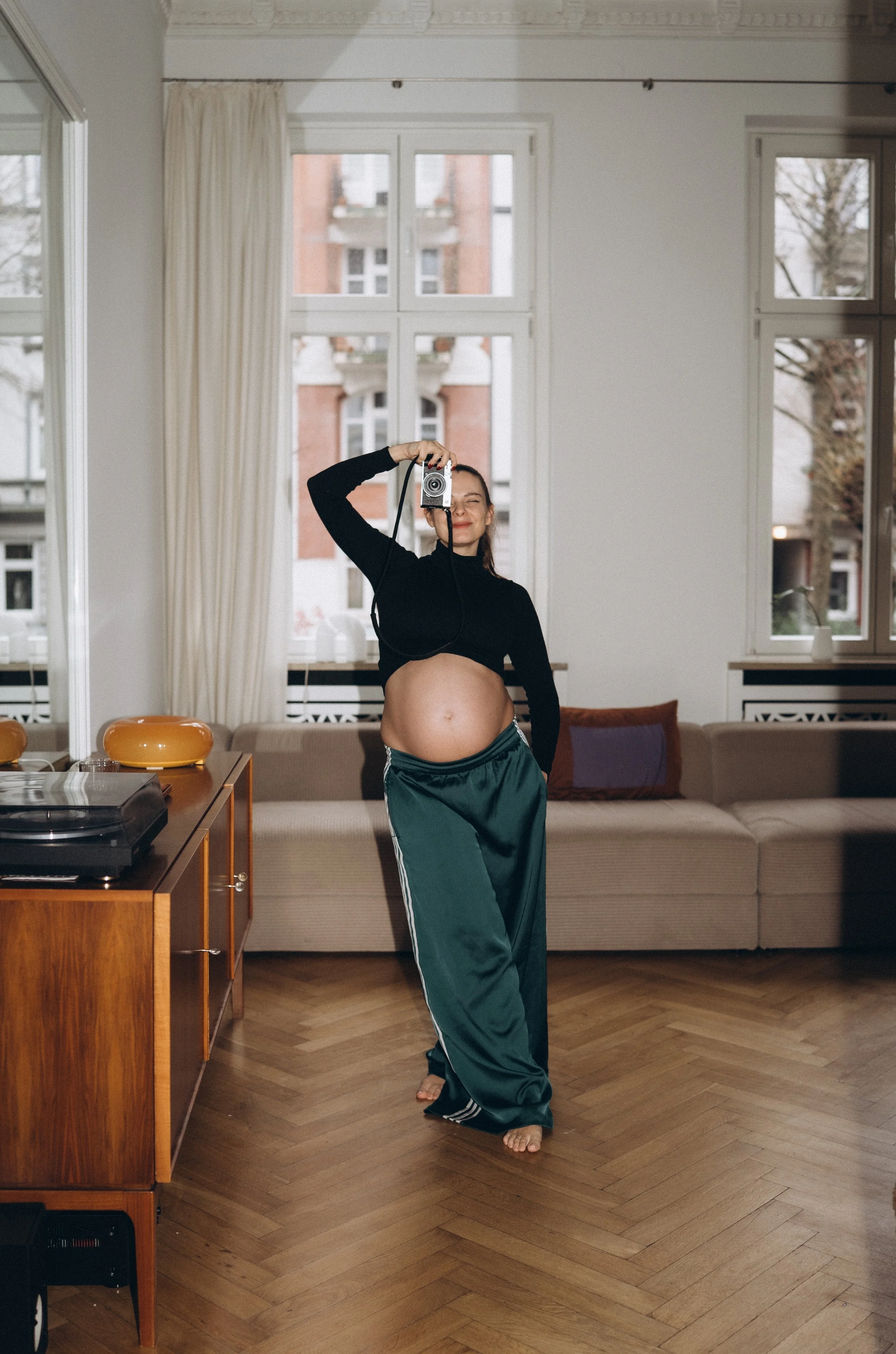 Schwangere Frau macht Selfie in einem Wohnzimmer vor einem Sofa, trägt schwarze Oberteil und grüne Jogginghose, hält Kamera in der Hand.