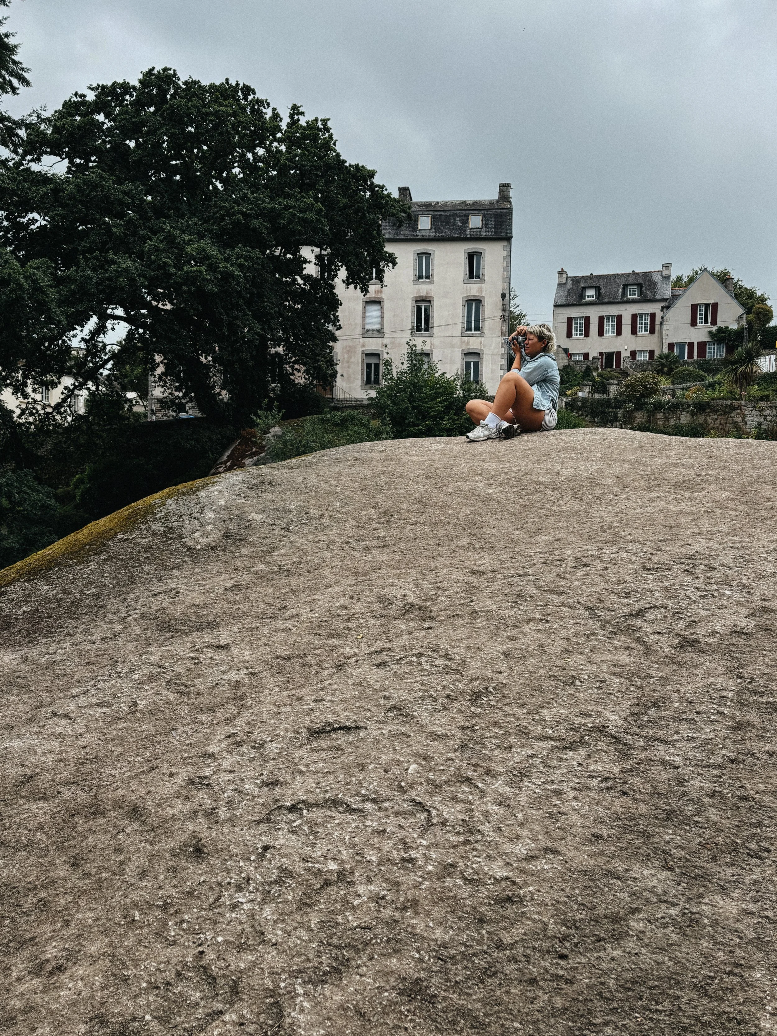 Junge Frau sitzt auf einem großen Felsen, fotografiert oder betrachtet die umliegende Stadt mit alten Gebäuden und Bäumen bei bewölktem Himmel.