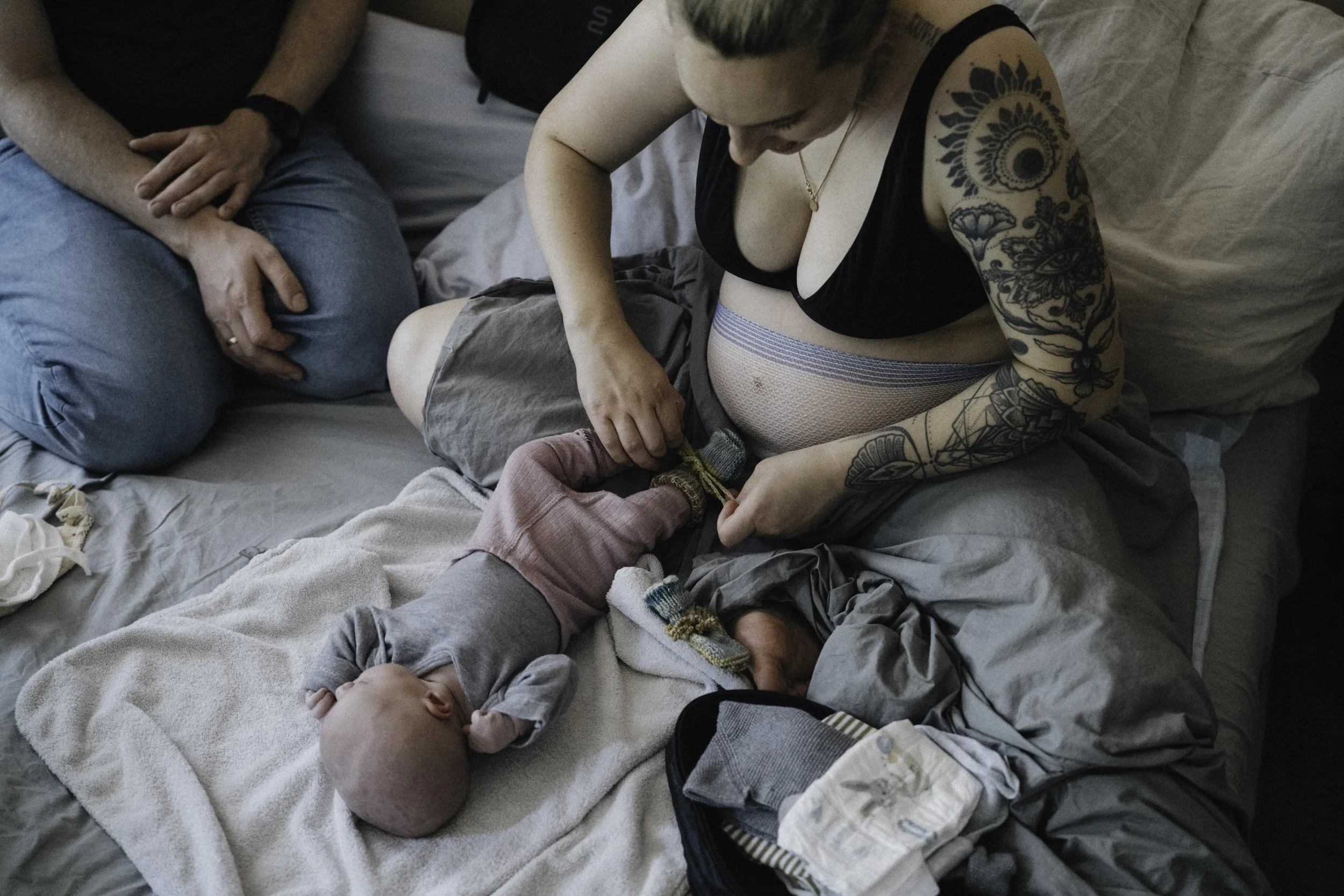 Eine Frau mit Tattoos, die ein Neugeborenes auf dem Bett wickelt, während eine zweite Person auf der Seite sitzt.