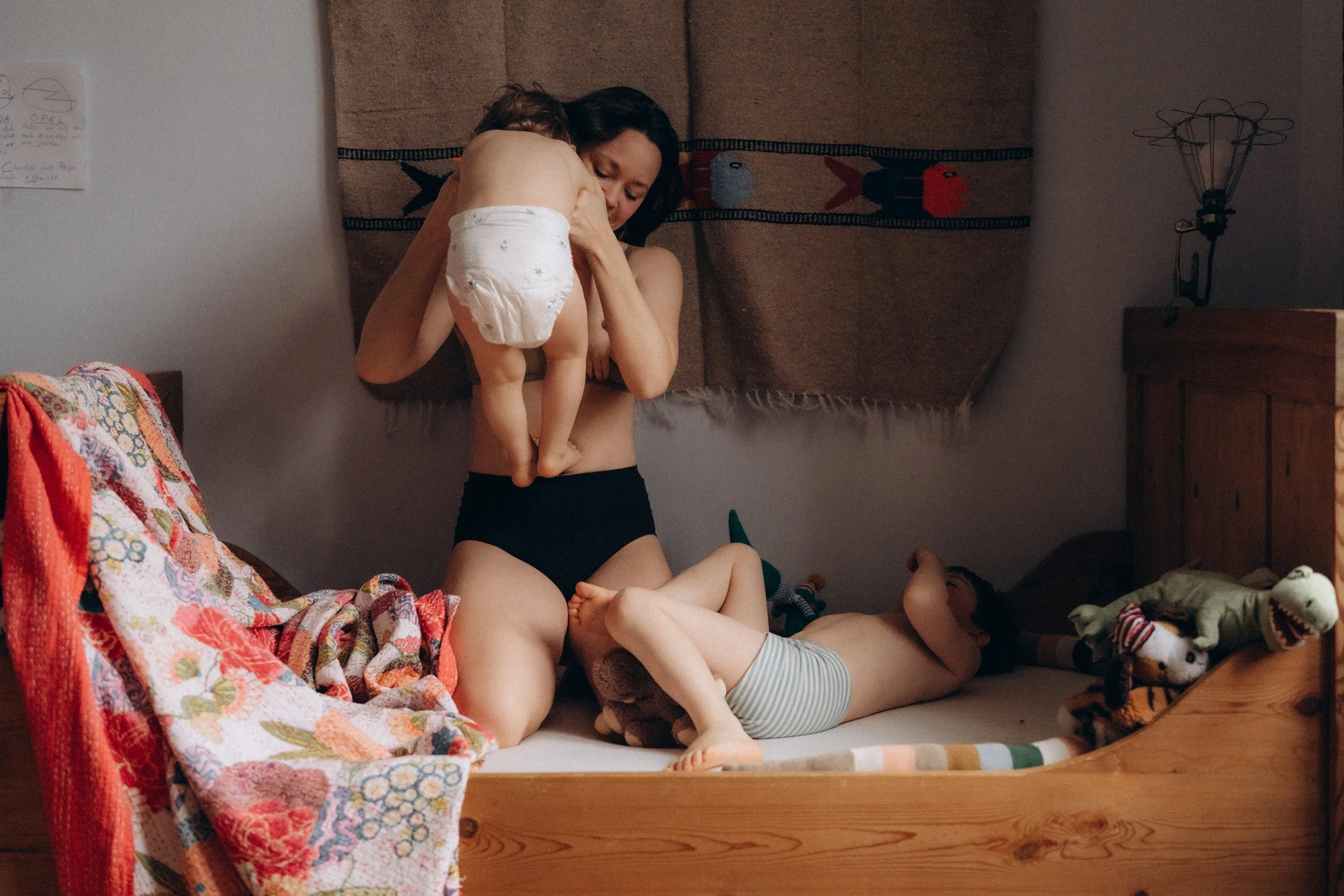 Eine Frau mit zwei kleinen Kindern in einem Schlafzimmer, eines der Kinder liegt auf dem Bett und das andere wird von der Frau gehalten, während sie spielen oder kuscheln.
