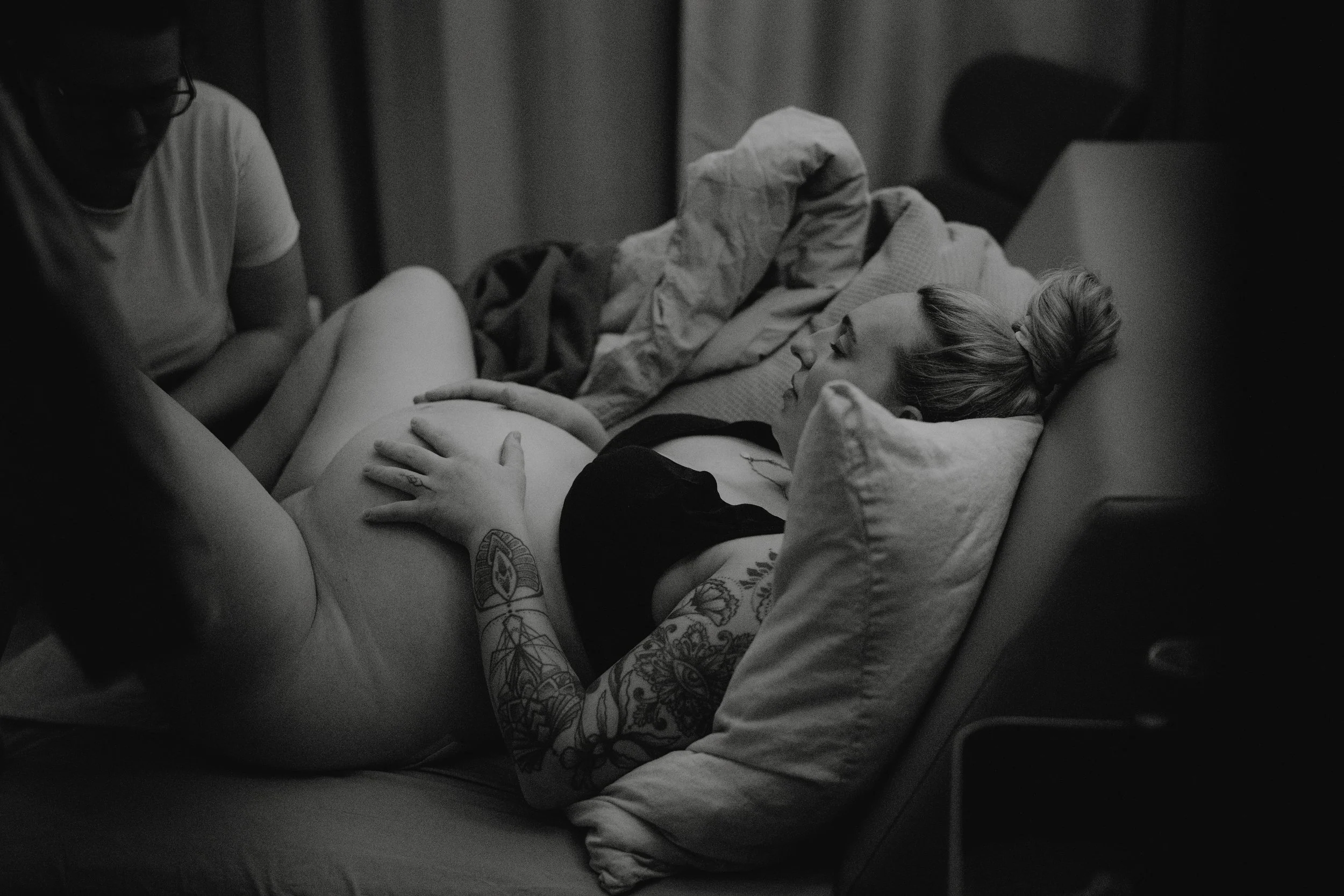 Eine schwangere Frau liegt auf einer Couch, von einer anderen Person begleitet. Es ist eine schwarze und weiße Aufnahme, die Entspannung und Nähe zeigt.