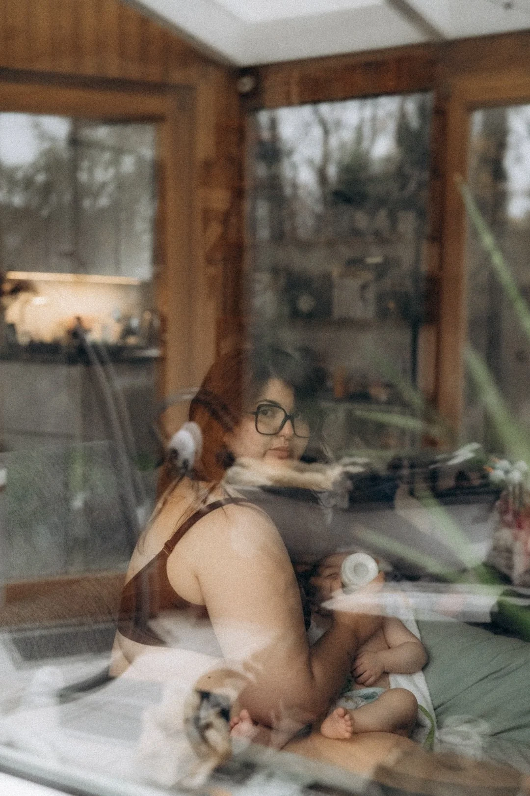 Ein Spiegelbild einer Frau mit schwarzen Haaren und Brille, die durch eine Glasscheibe blickt, während sie ein Baby auf dem Schoß hält, im Hintergrund ein Raum mit Holzrahmen und Fenstern, draußen Bäume sichtbar.