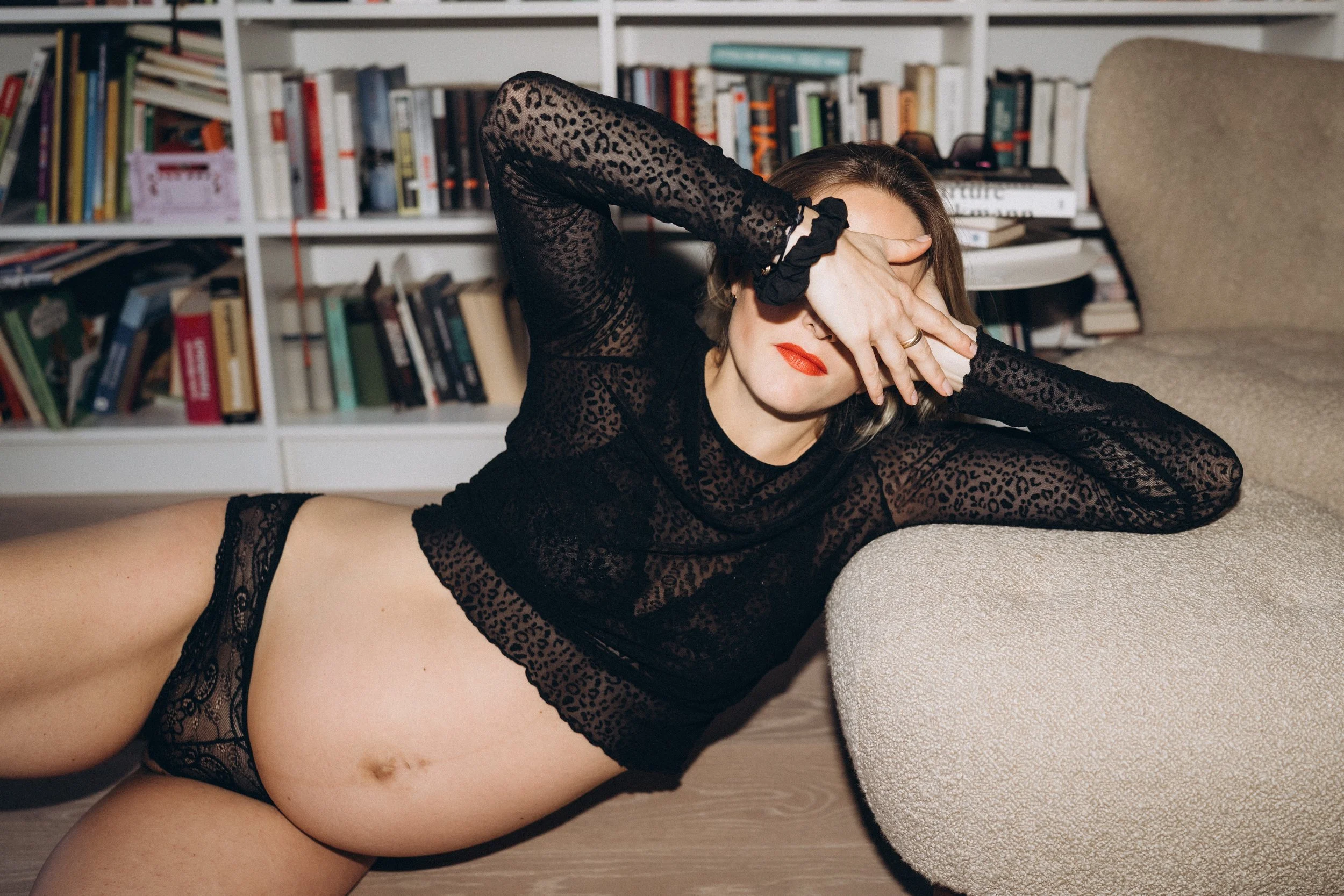 Frau in schwarzem Spitzenkleid und Unterwäsche liegt auf dem Boden vor einem Bücherregal, mit einer Hand vor dem Gesicht.