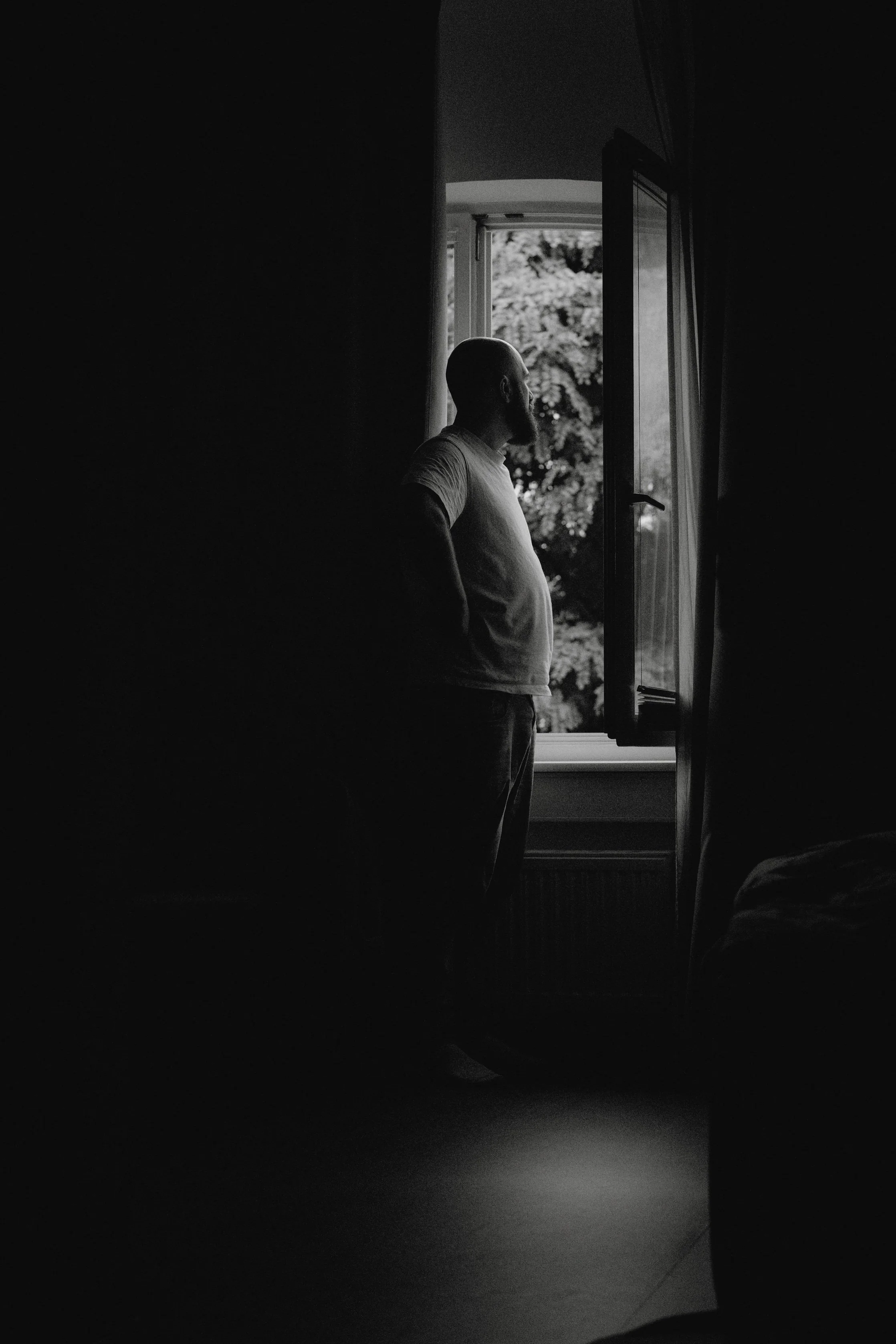 Ein Mann steht am offenen Fenster eines Zimmers und blickt nach draußen, umgeben von natürlichem Licht, das durch das Fenster scheint.