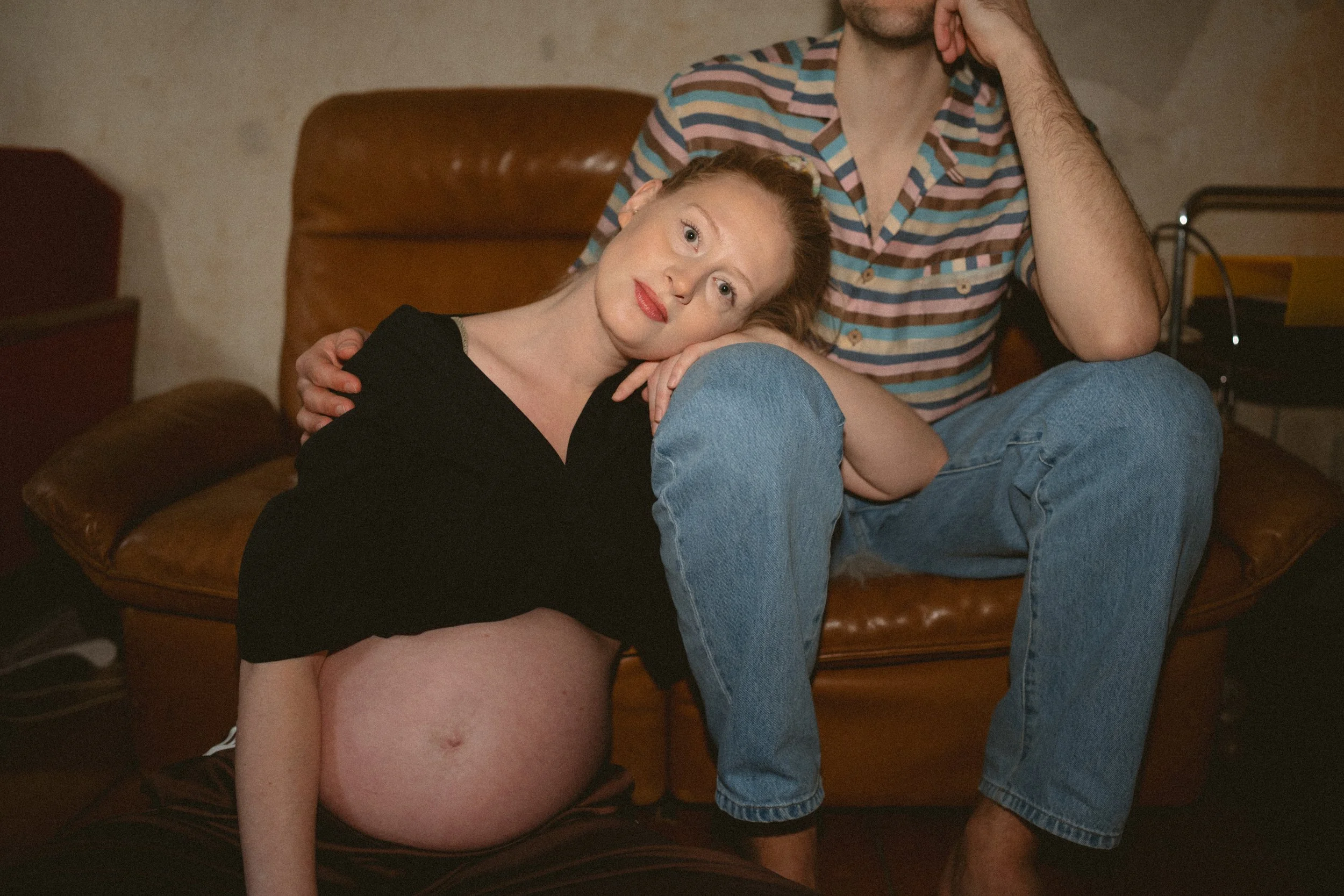 Ein schwangere Frau und ein Mann sitzen entspannt auf einem braunen Sofa im Raum, die Frau lehnt ihren Kopf an die Hand des Mannes, der ihn auf ihre Schulter stützt. Die Frau trägt ein schwarzes Oberteil, das ihren wachsenden Bauch zeigt, während der Mann ein bunt gestreiftes Hemd und blaue Jeans trägt.