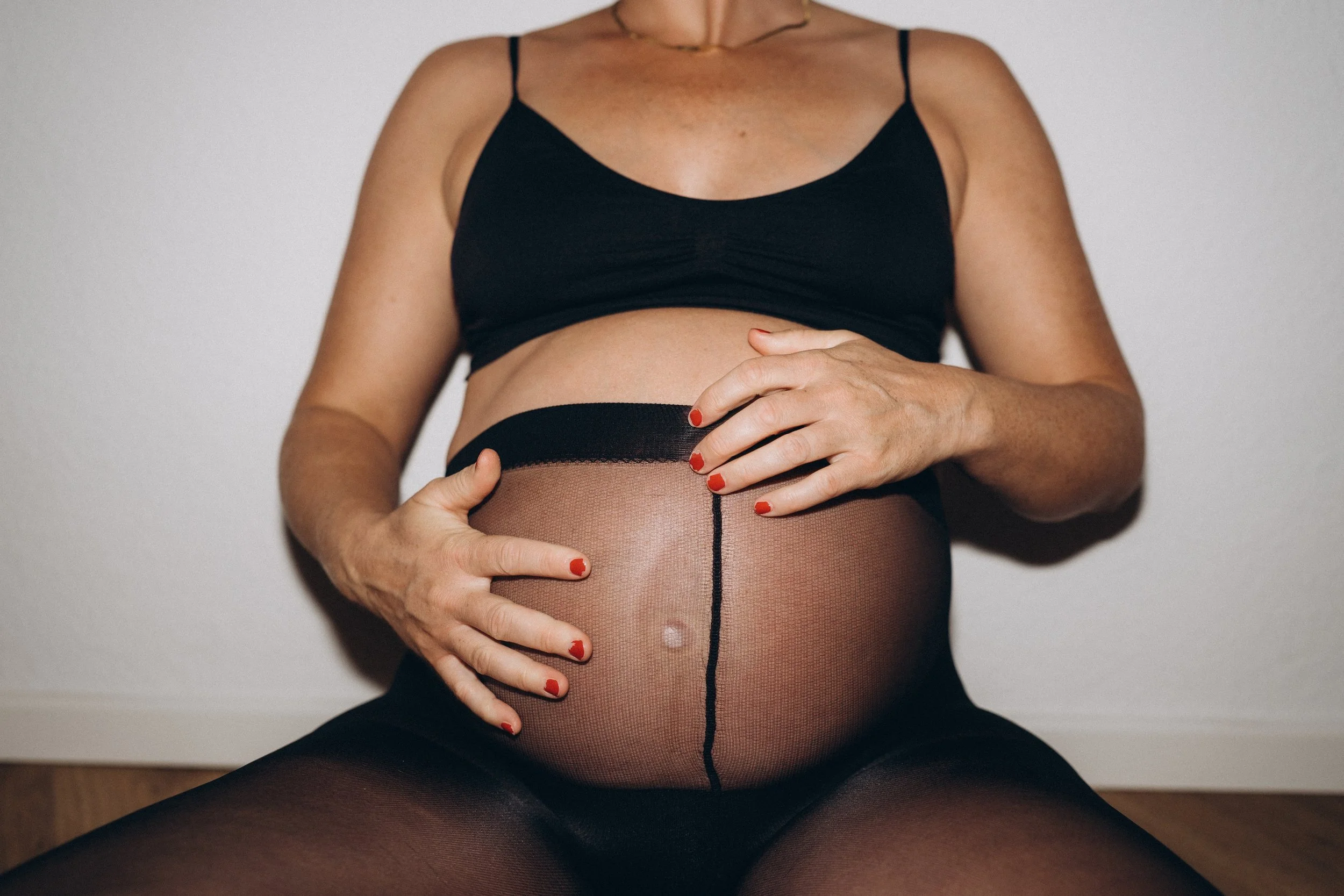 Eine schwangerer Frau in schwarzer Unterwäsche und Netzstrümpfen sitzt auf dem Boden, hält ihren Babybauch mit beiden Händen und hat rote Nagellackfarben.